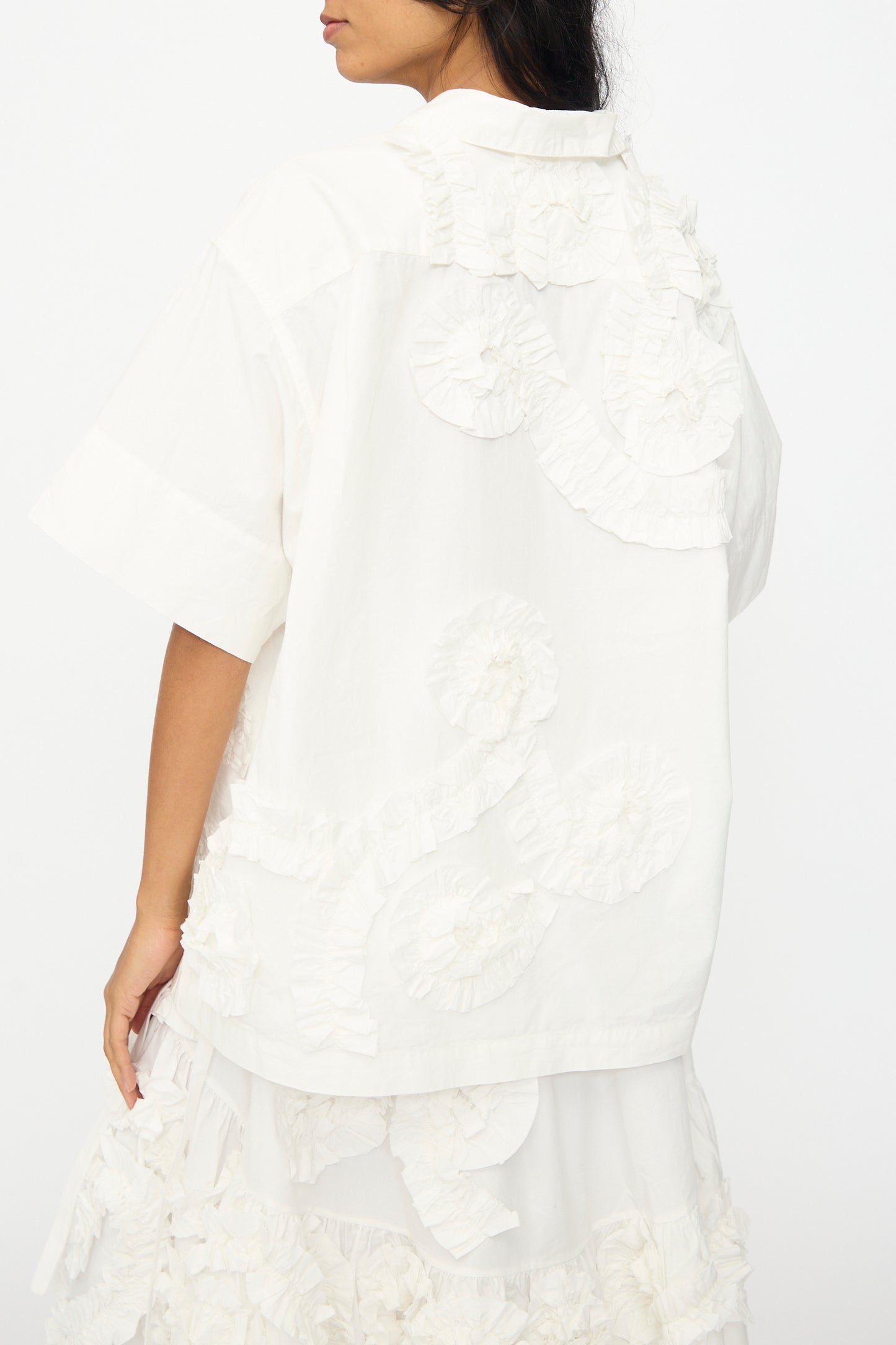 A person wearing the Story mfg. Hand Applique Greetings Shirt in Ecru Fossil, an oversized short-sleeve shirt, is shown from the back and side against a plain white background.