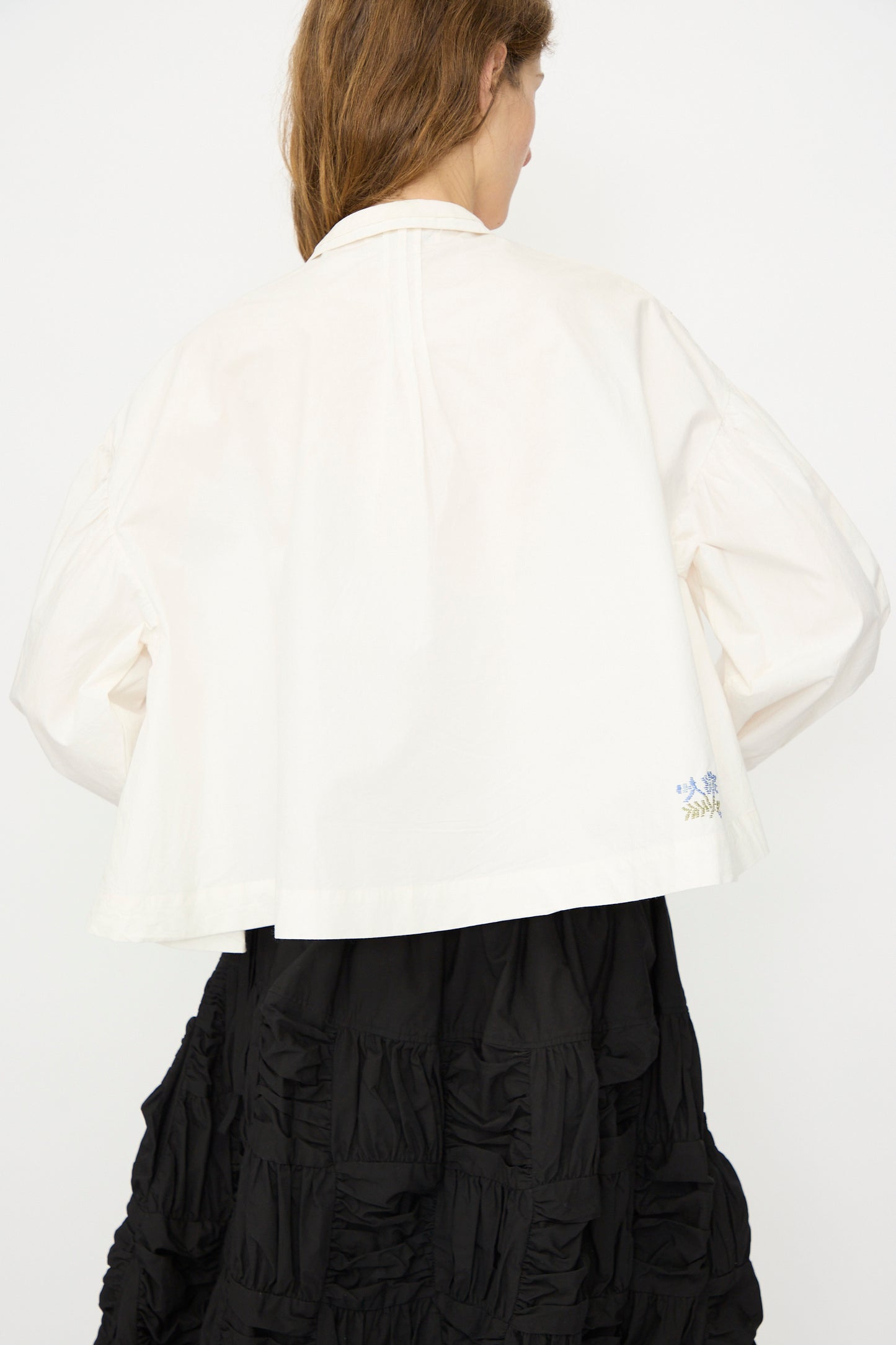 A person facing away wears the Story mfg. Still Shirt in Interrupted Embroidery—an organic cotton blouse with subtle blue hand embroidery near the hem—paired with a black textured skirt, set against a plain white background.