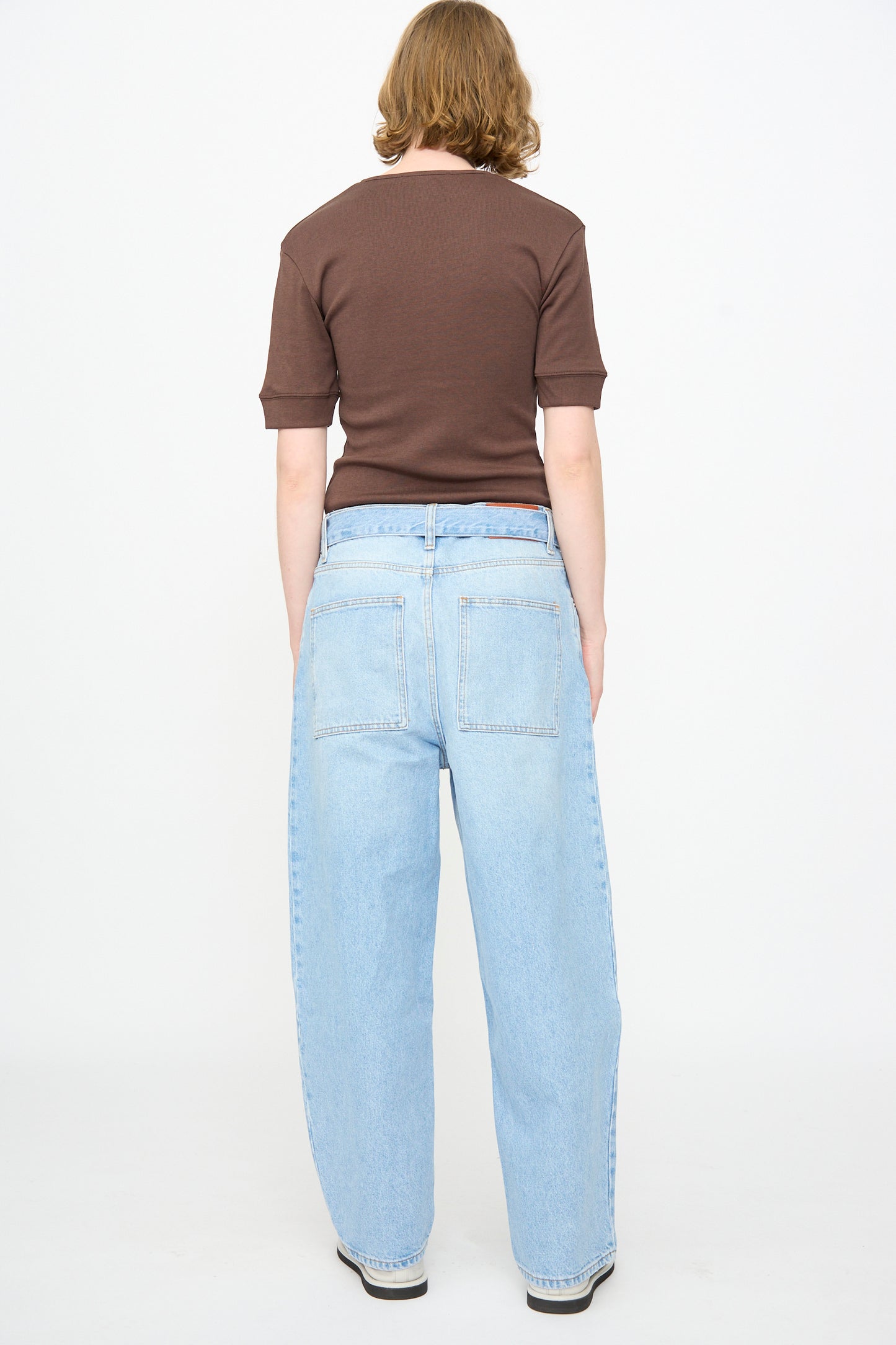 A person faces away from the camera, wearing a short-sleeve brown shirt and Studio Nicholson 14 oz. Denim Cahun Pant in Bleached Wash—light blue, wide-leg relaxed jeans—while standing against a plain white background.