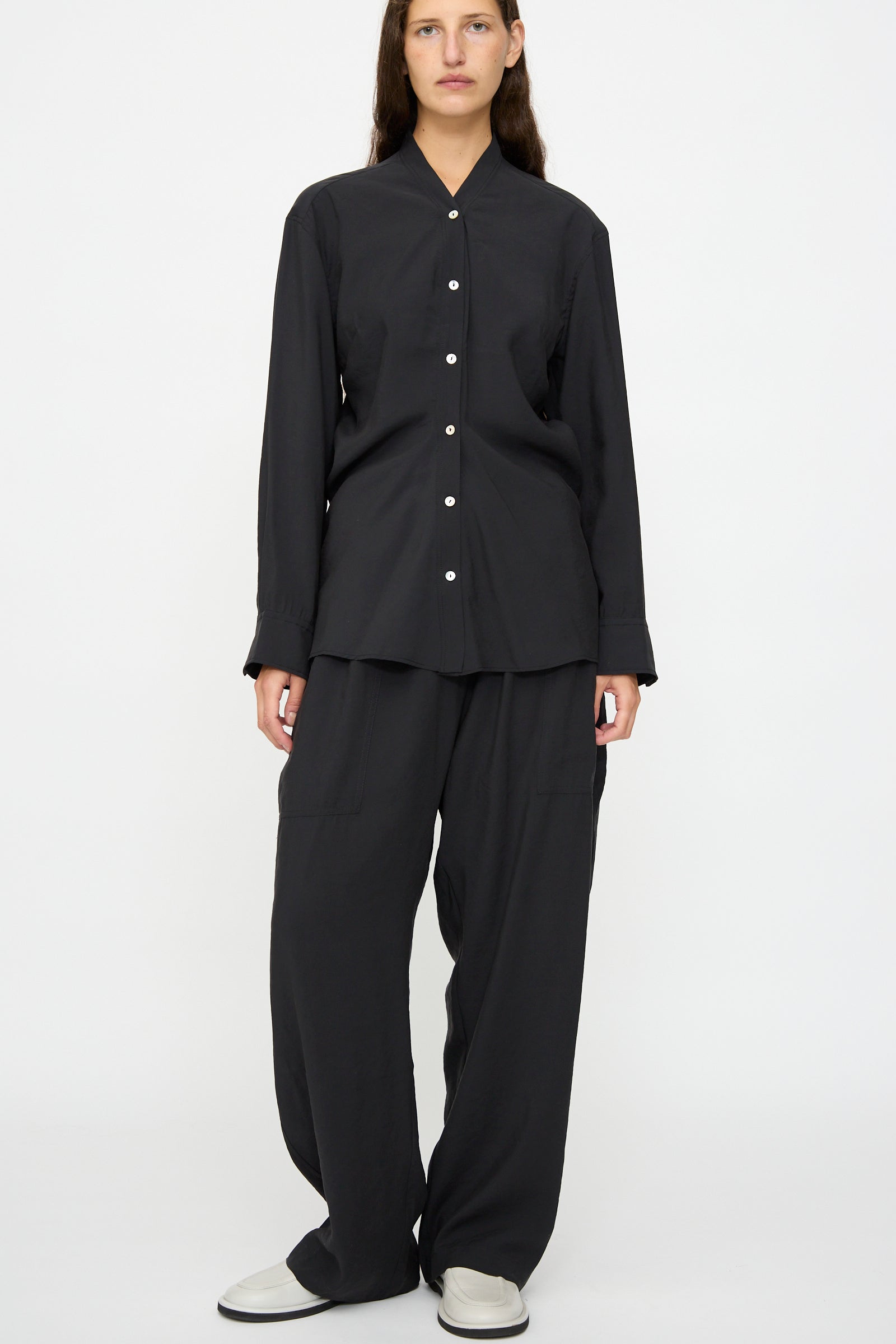 A person stands facing forward against a plain white background, wearing the Studio Nicholson Acre Top in Darkest Navy—a loose-fit, long-sleeve collarless button-up shirt—paired with wide black pants and white shoes.