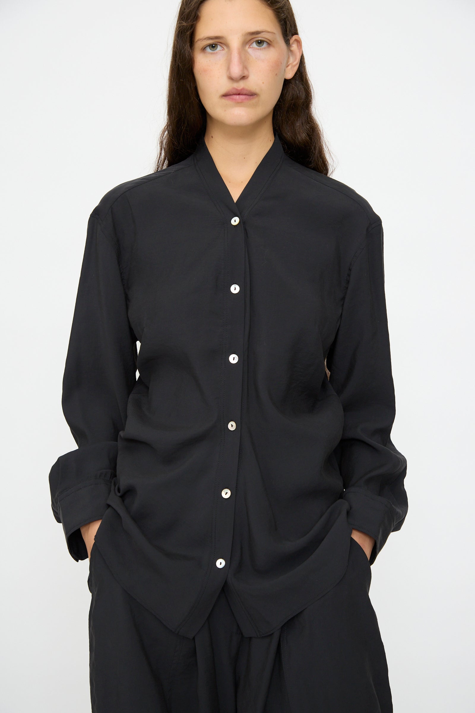 A person with long brown hair wears the Studio Nicholson Acre Top in Darkest Navy, a loose-fit, collarless button-up shirt, paired with matching black pants, standing against a plain white background.