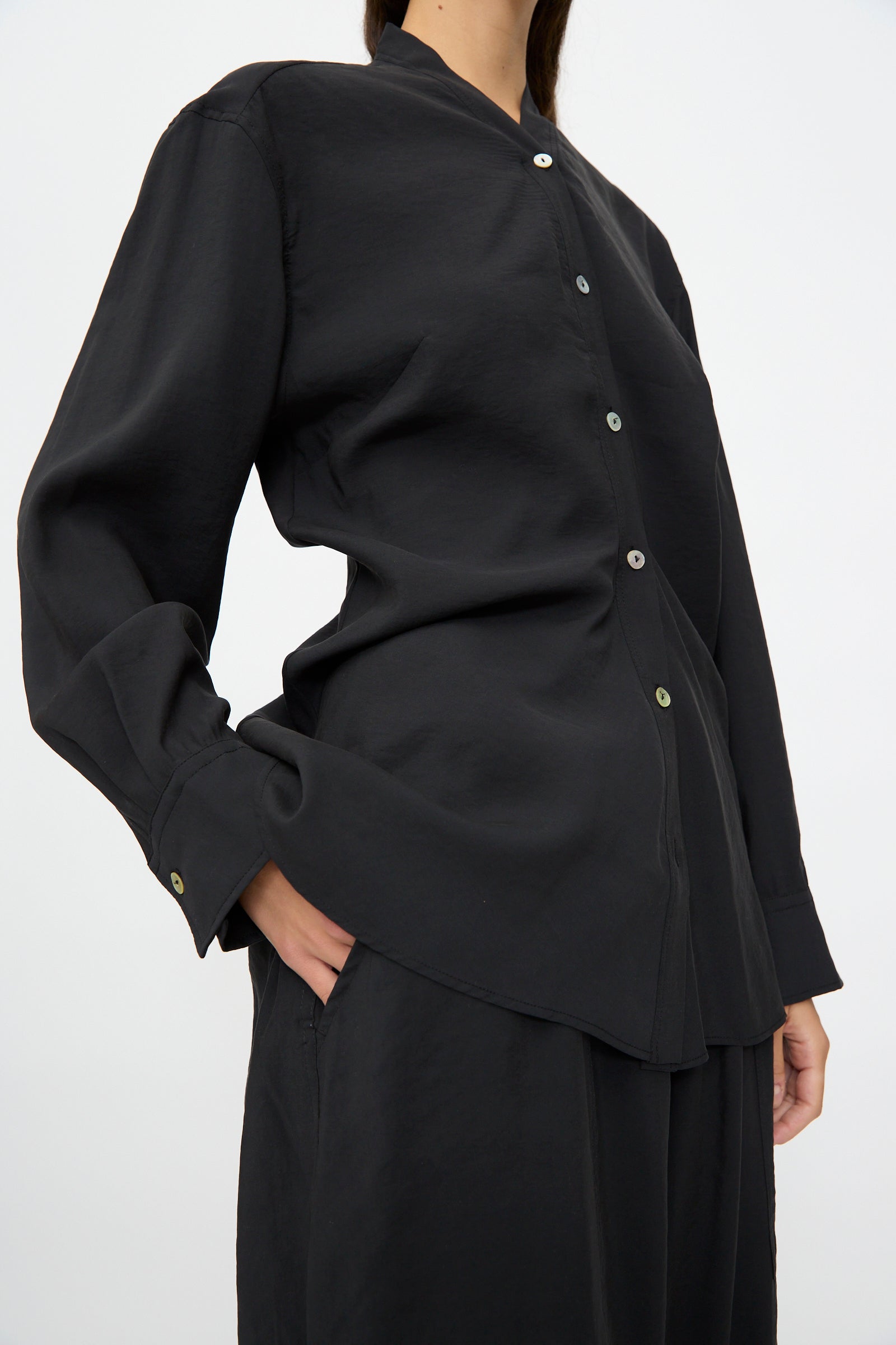 Person wears the Studio Nicholson Acre Top in Darkest Navy, a loose fit, collarless button-up shirt, paired with matching black pants, one hand in their pocket, against a plain light background.