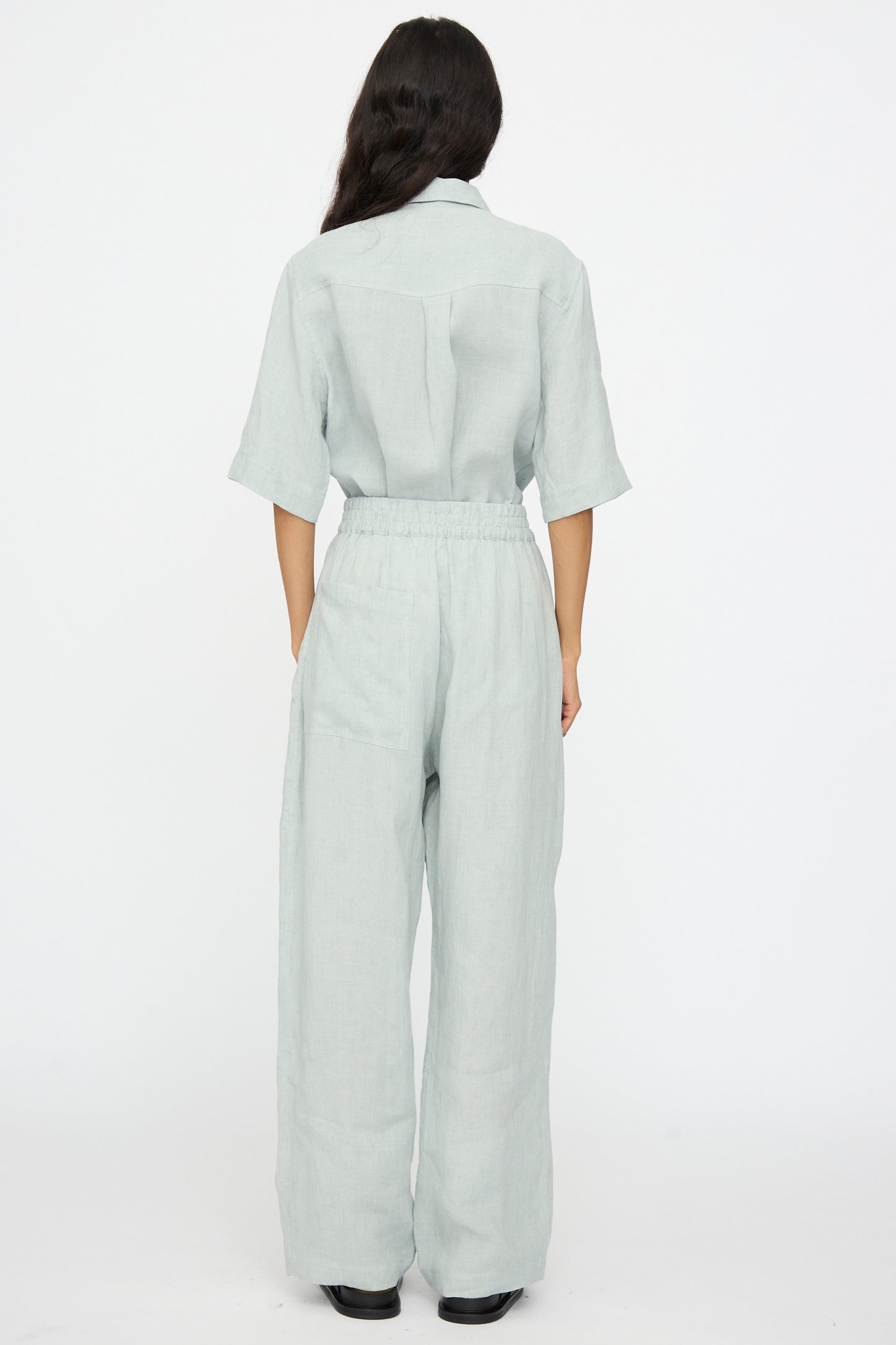 A person stands with their back to the camera, wearing Studio Nicholson’s Attis Pant in Spearmint—light-colored, loose-fitting with an elastic waistband and wide legs—paired with black shoes against a plain white background.