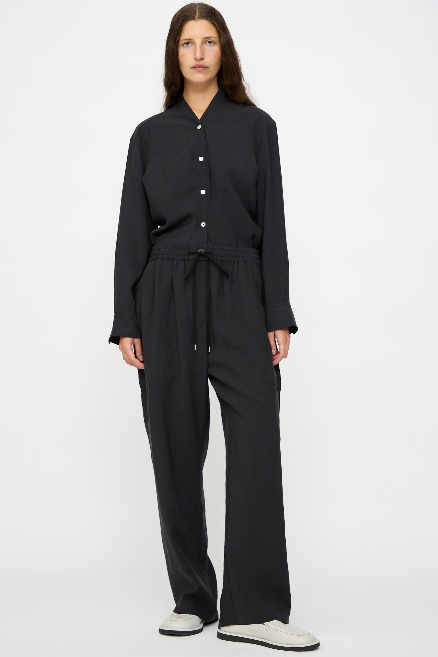 A person faces forward wearing Studio Nicholson’s Capel Pant in Darkest Navy with a relaxed long-sleeve black button-up shirt and white slip-on shoes, standing against a plain background.