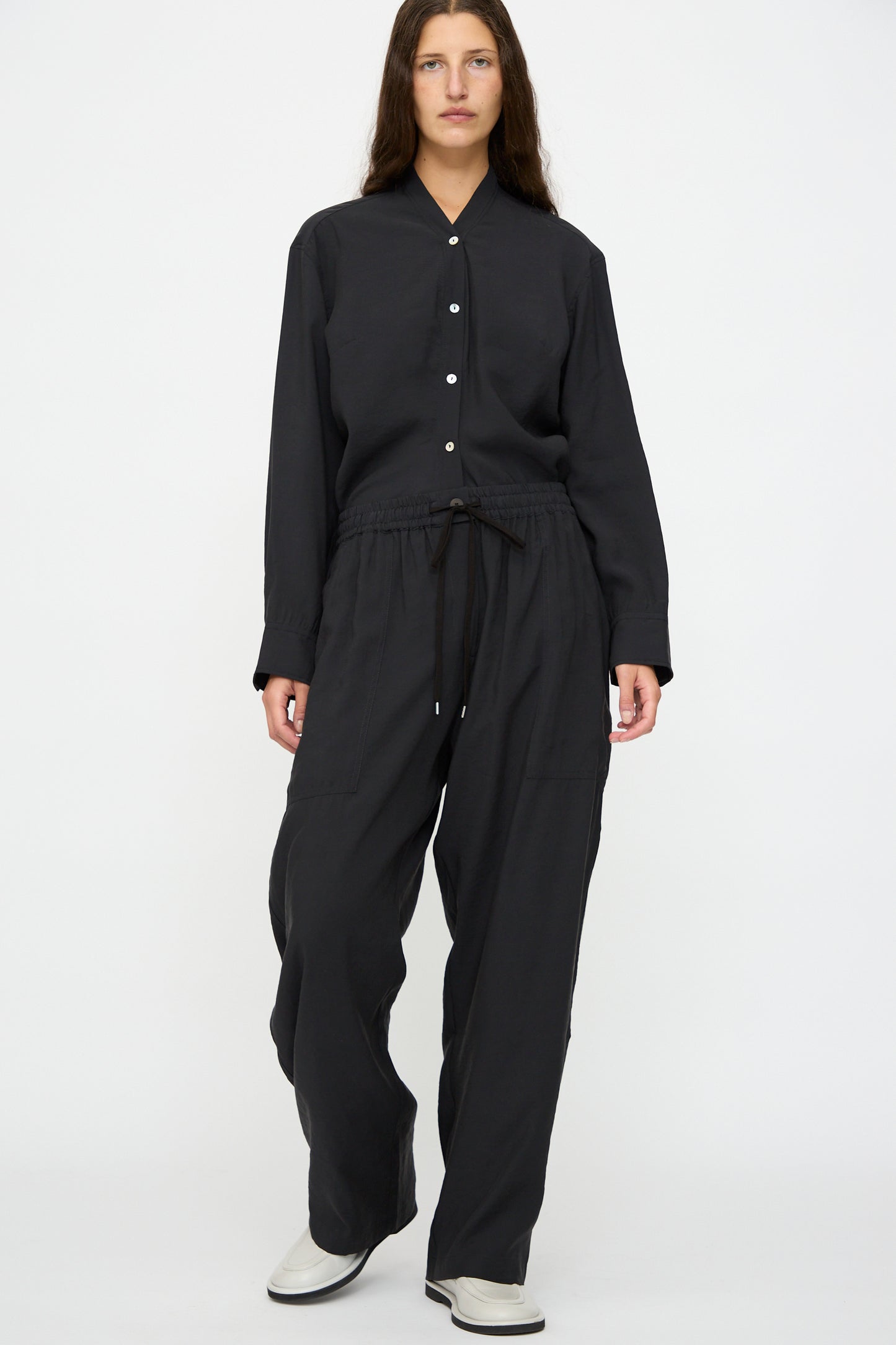 A person stands facing forward, wearing Studio Nicholson's Capel Pant in Darkest Navy, styled with a relaxed fit black button-up shirt and white shoes, against a plain white background.