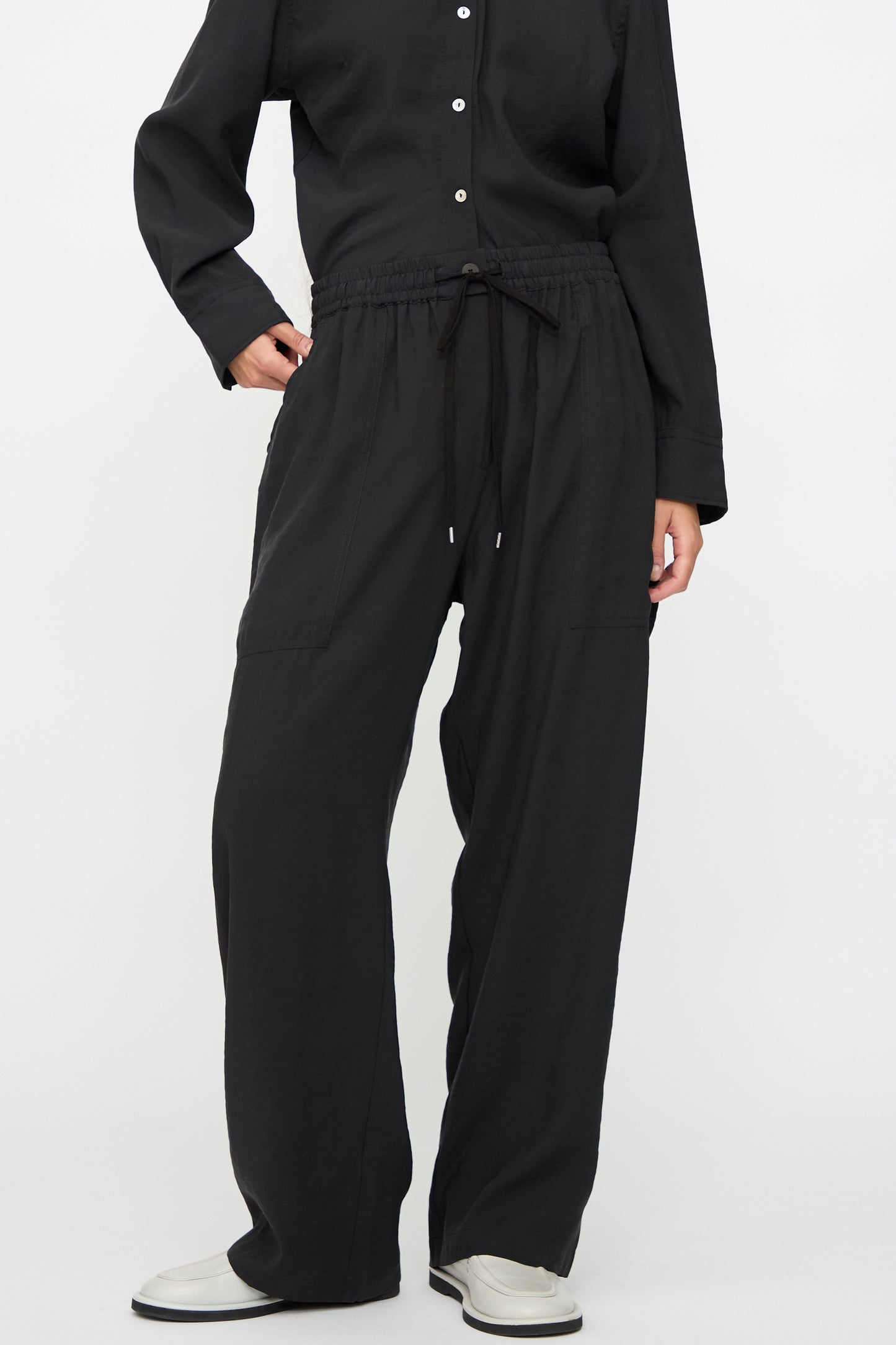 A person wears a long-sleeve black button-up shirt, Studio Nicholson Capel Pant in Darkest Navy (relaxed fit, modal blend), and white shoes, standing against a plain white background.