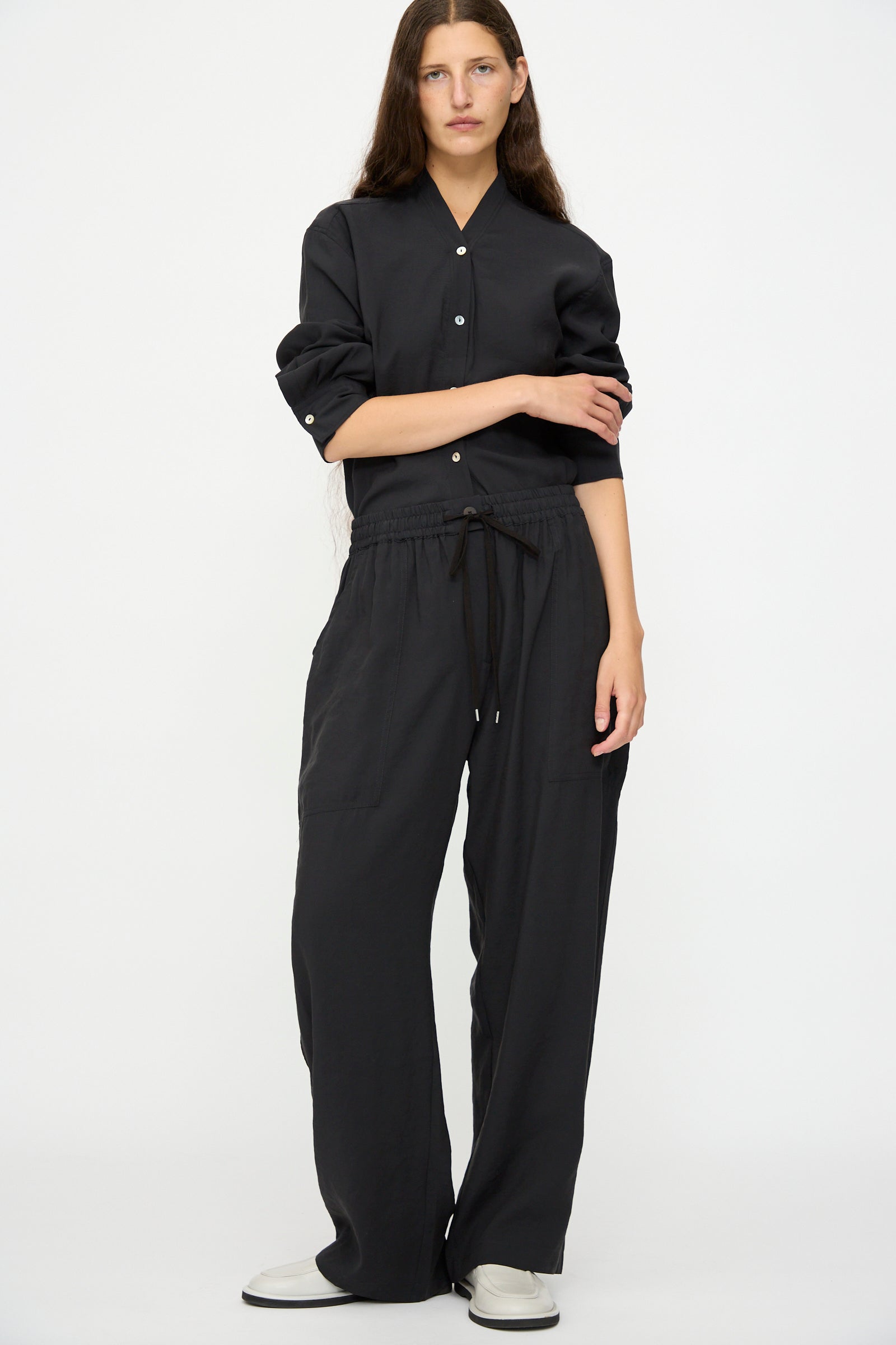 A person stands against a plain background wearing Studio Nicholson's Capel Pant in Darkest Navy with a relaxed fit black button-up shirt and white shoes.