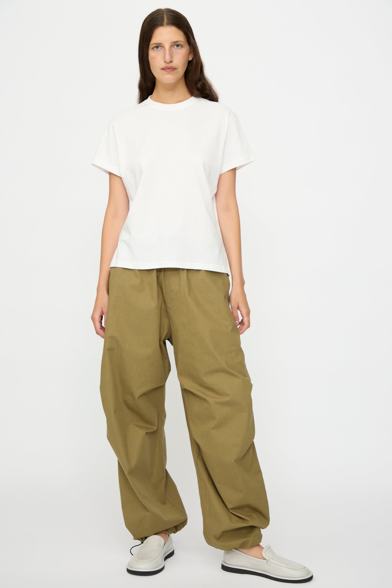 A person faces forward against a white background, wearing Studio Nicholson's Caro Belted Pant in Military Green, a plain white t-shirt, and white slip-on shoes.