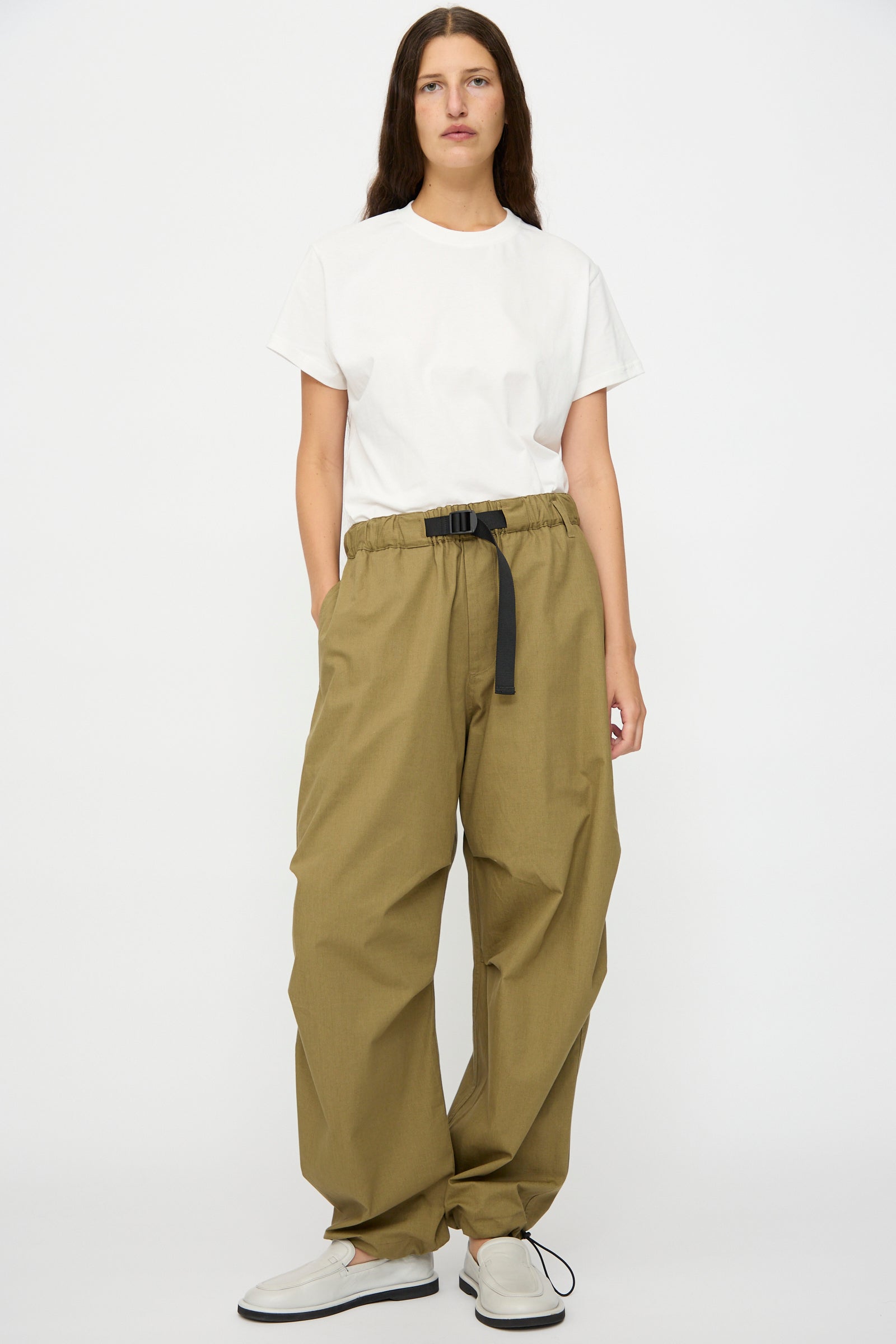 A person stands against a white background wearing a plain white t-shirt, Studio Nicholson Caro Belted Pant in Military Green with a black belt, and white slip-on shoes.
