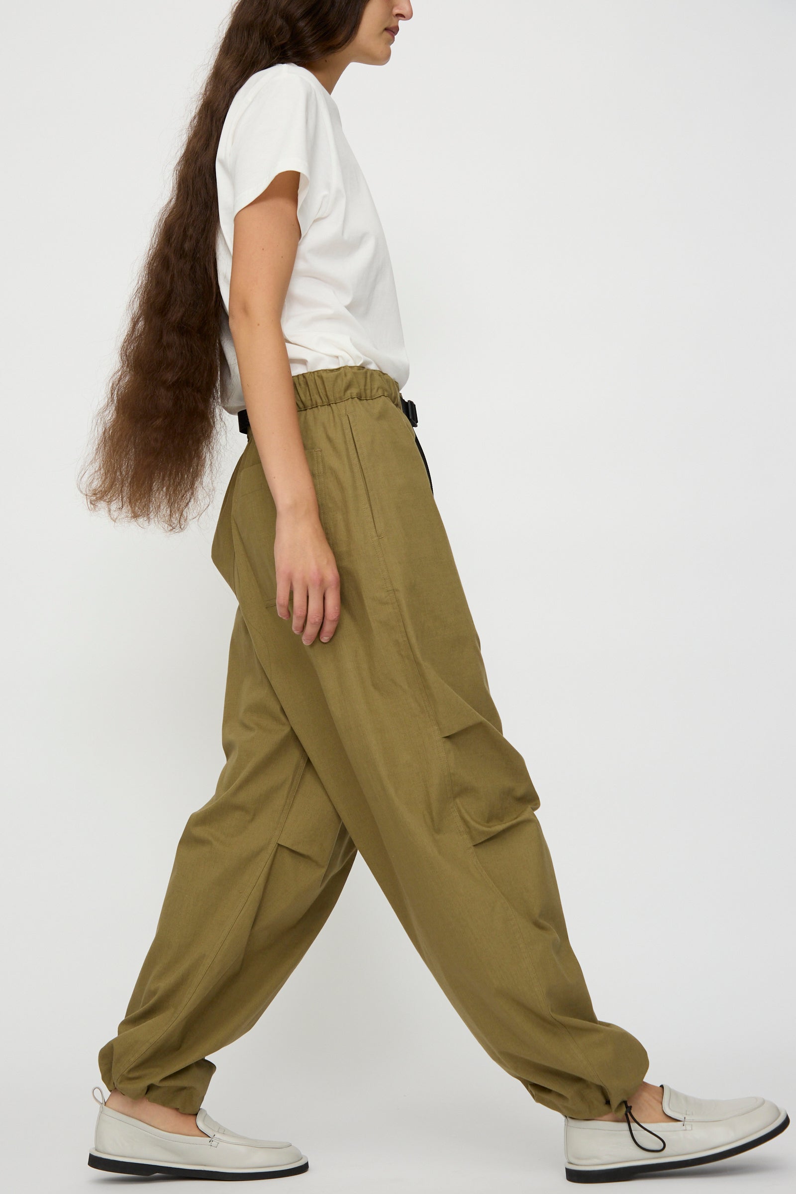 A person with long brown hair wears a white t-shirt, Studio Nicholson Caro Belted Pant in Military Green, and white slip-on shoes, walking against a plain white background.