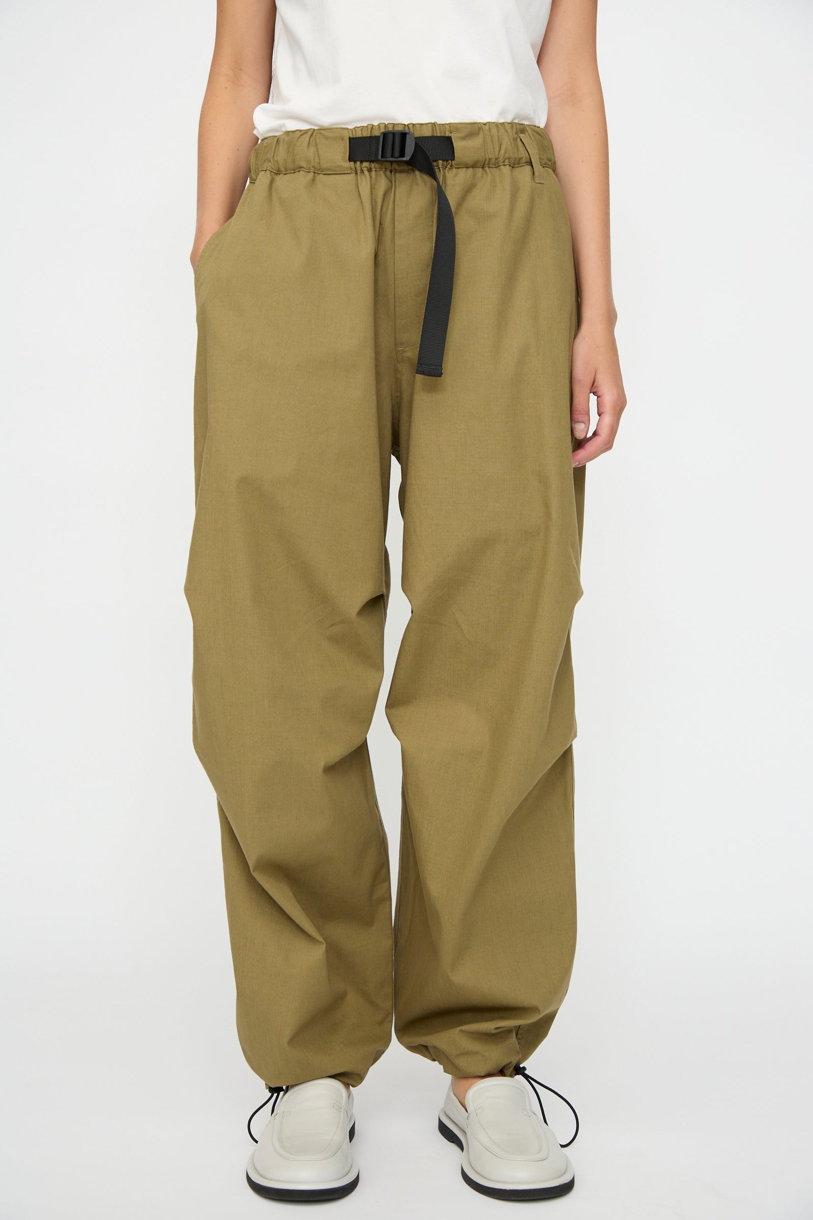 A person wears the Studio Nicholson Caro Belted Pant in Military Green with a black belt, white shirt, and white shoes, standing against a plain white background.