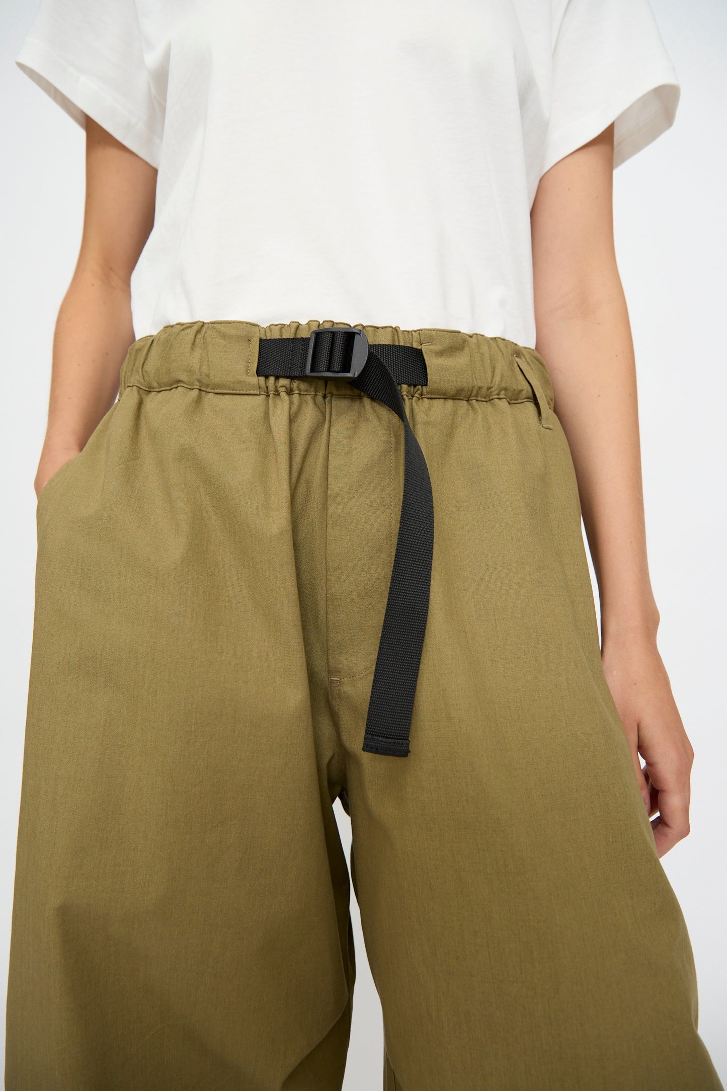 A person wears the Studio Nicholson Caro Belted Pant in Military Green, styled with a white t-shirt and adjustable black belt, hands in pockets.