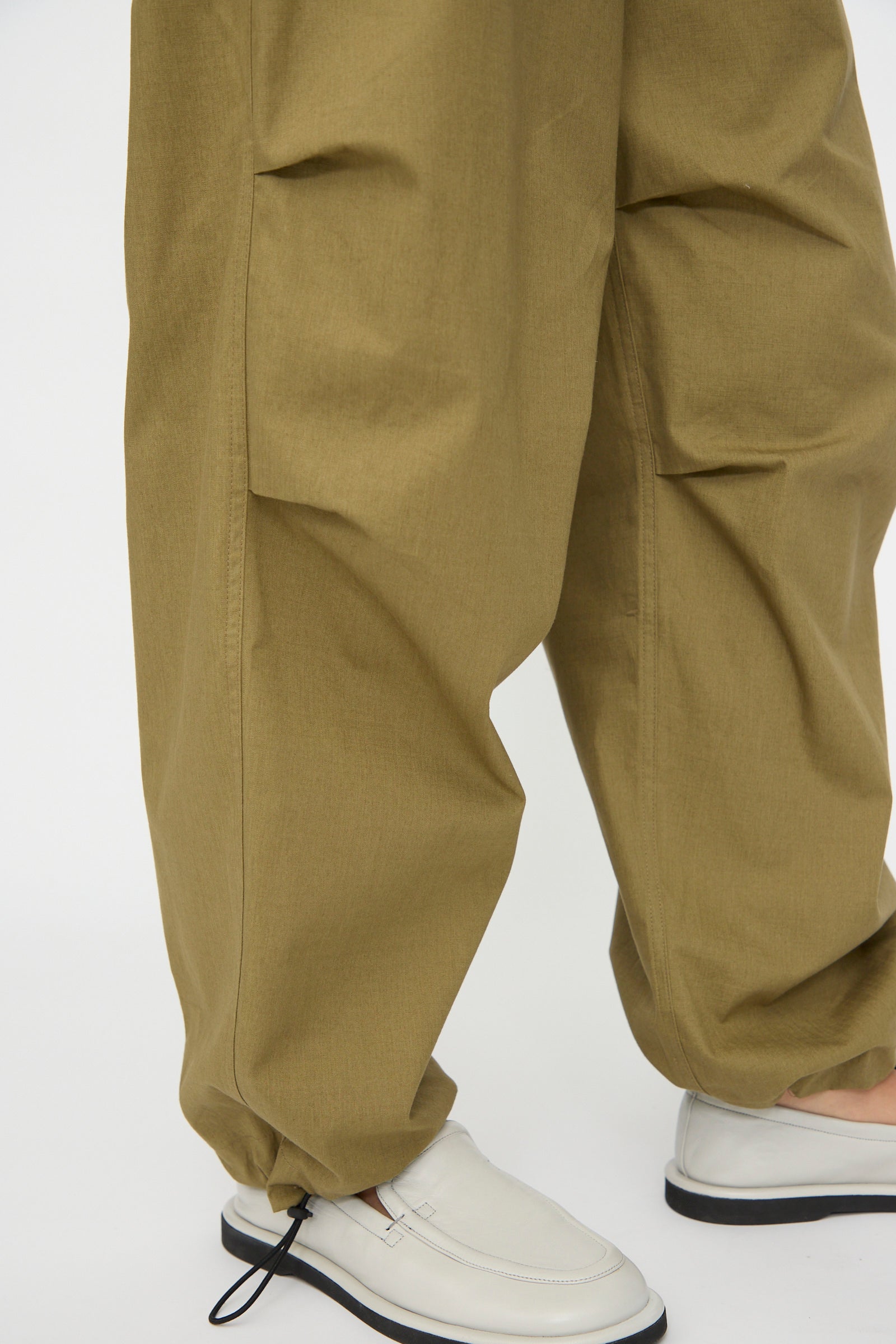 A person wearing Studio Nicholson's Caro Belted Pant in Military Green with drawstring cuffs and white slip-on shoes, standing on a white background.