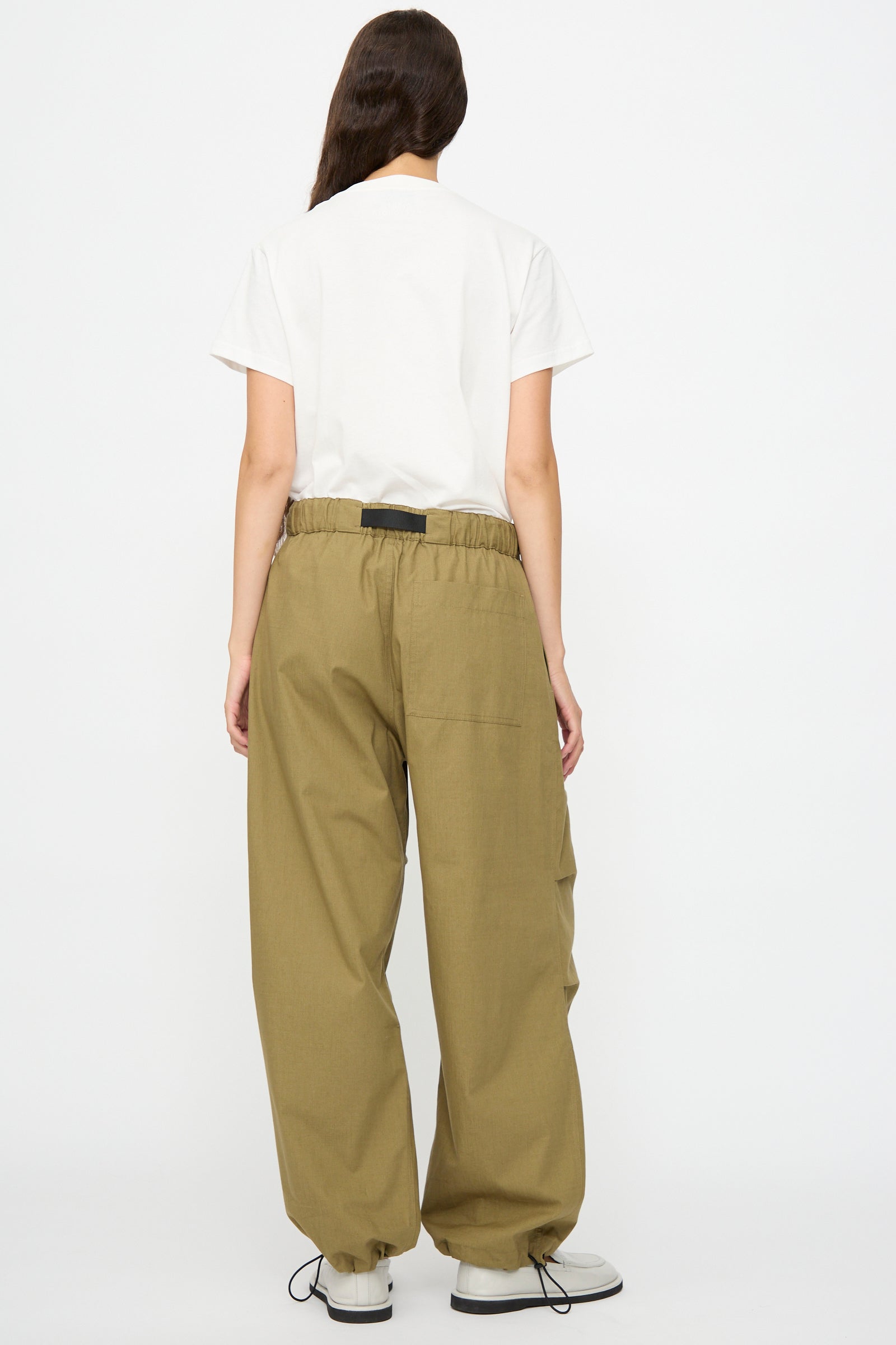 A person faces away, wearing Studio Nicholson's Caro Belted Pant in Military Green with a white t-shirt and white shoes, set against a plain white background.