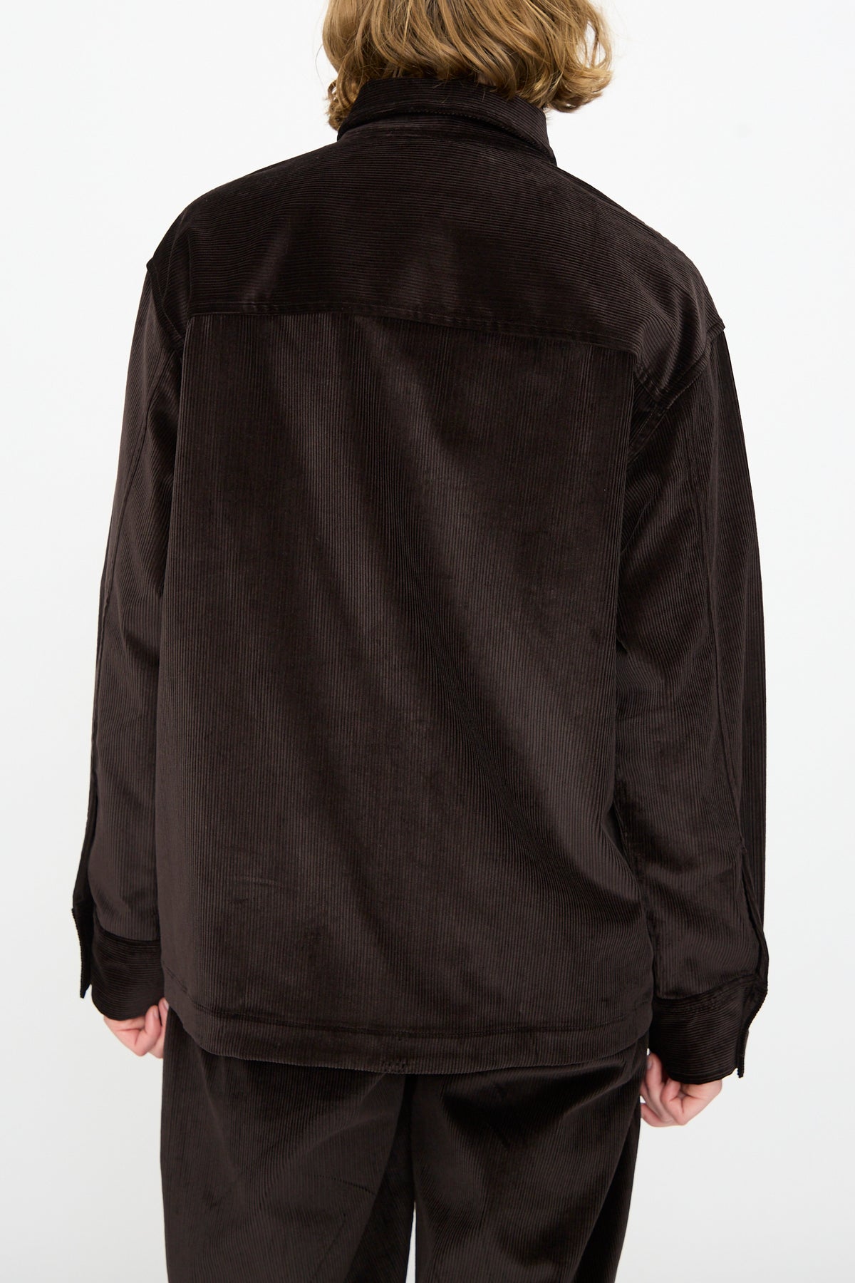 A person with medium-length hair, shown from the back against a plain white background, is wearing the Studio Nicholson Cotton Corduroy Santini Overshirt in Espresso paired with matching pants in a relaxed fit.