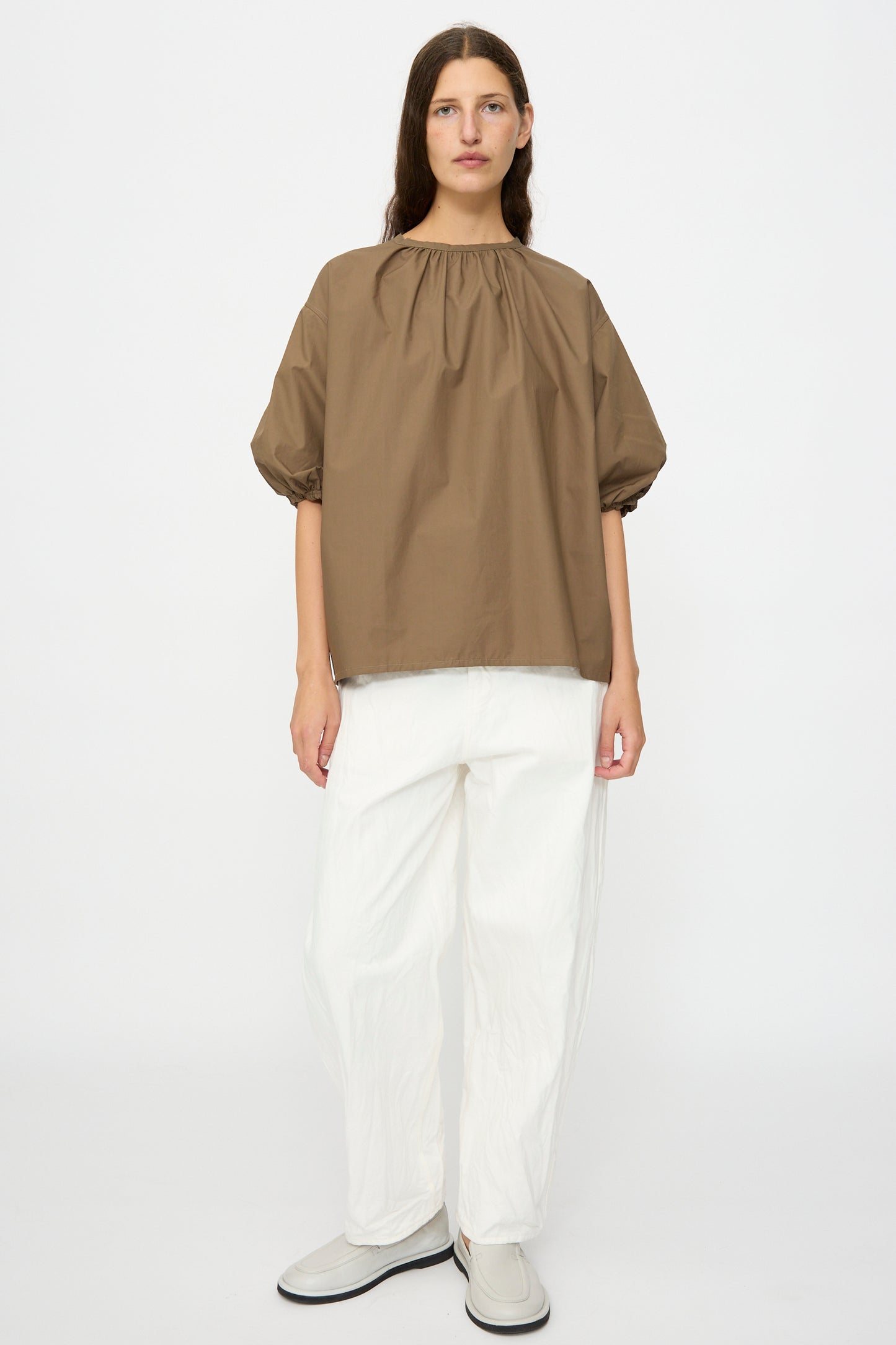 A person stands against a plain background wearing the Studio Nicholson Cove Gathered Top in Boulder, paired with white wide-leg pants and light-colored slip-on shoes.
