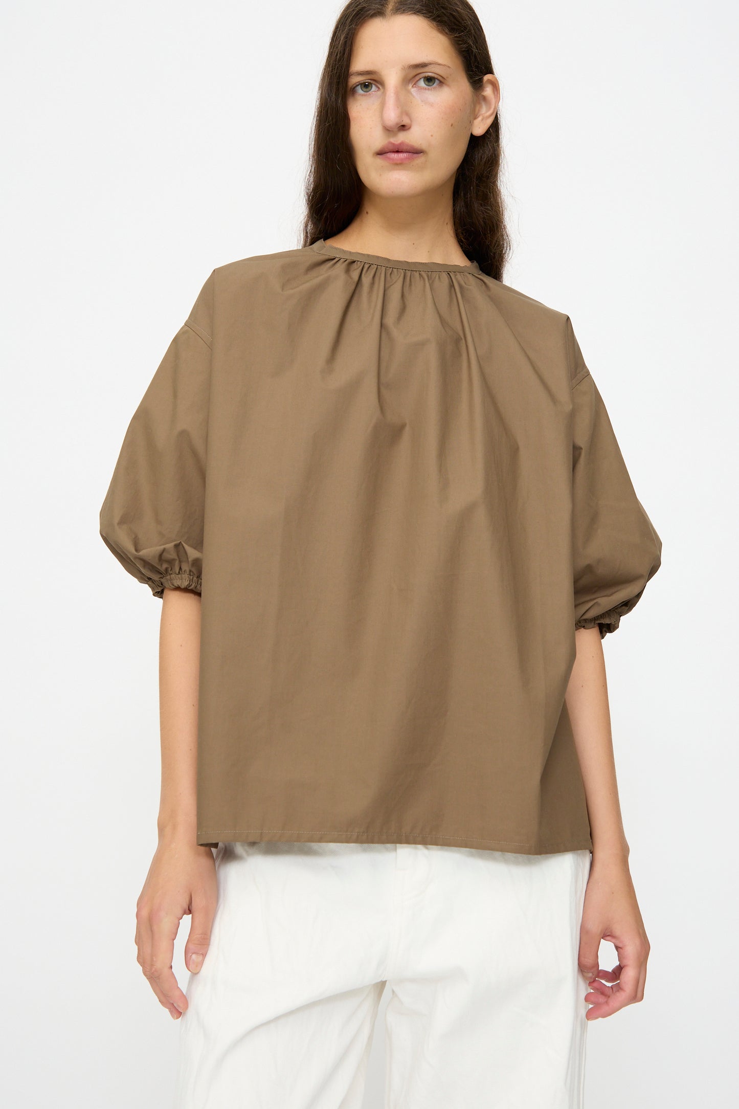 A person wearing the Studio Nicholson Cove Gathered Top in Boulder, a relaxed fit cotton short sleeve with a gathered neckline, and white pants stands against a plain light background.