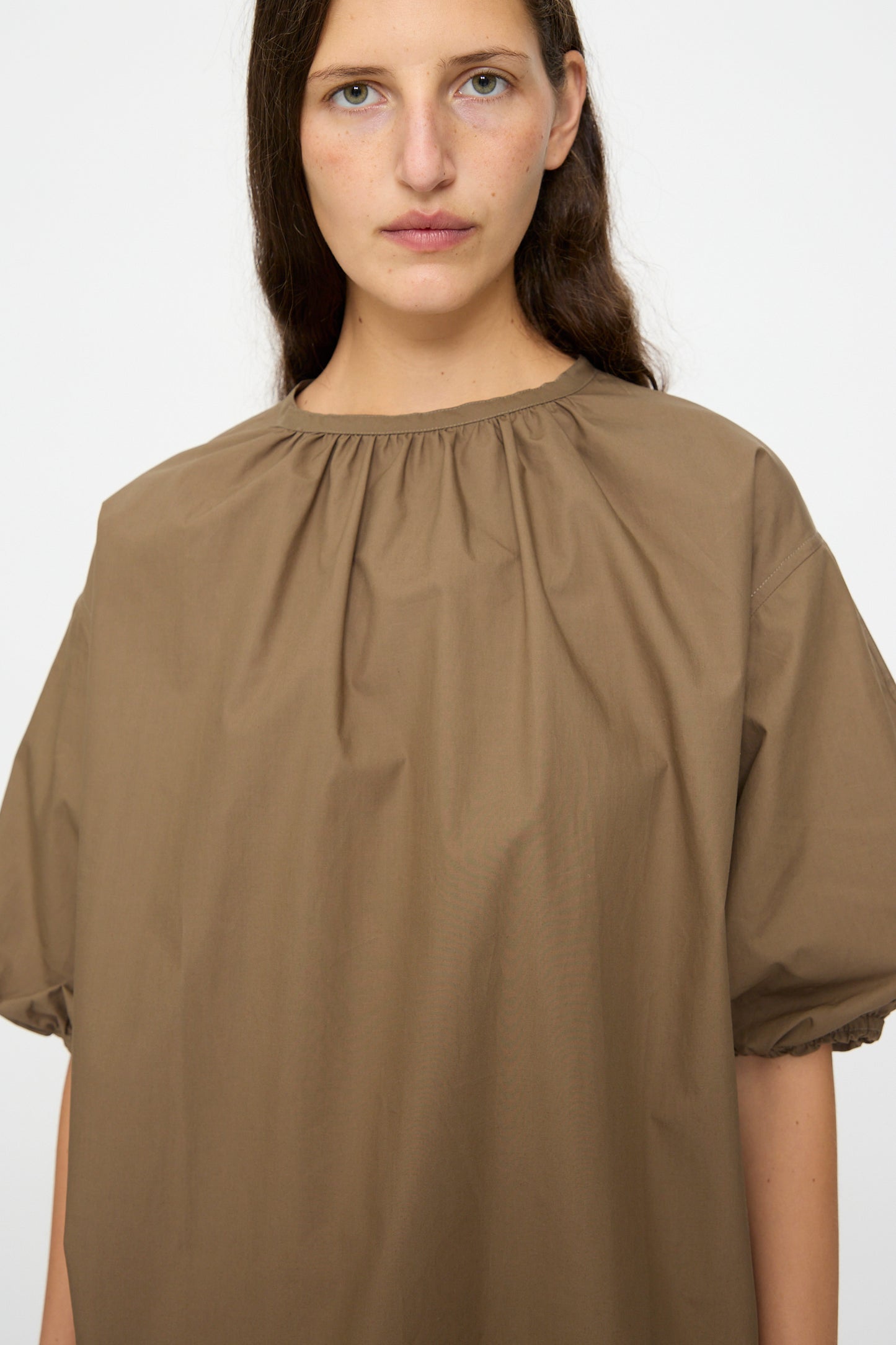 A person with long brown hair wears the Studio Nicholson Cove Gathered Top in Boulder, a relaxed-fit brown cotton short-sleeve style with a gathered neckline, standing against a plain light background.
