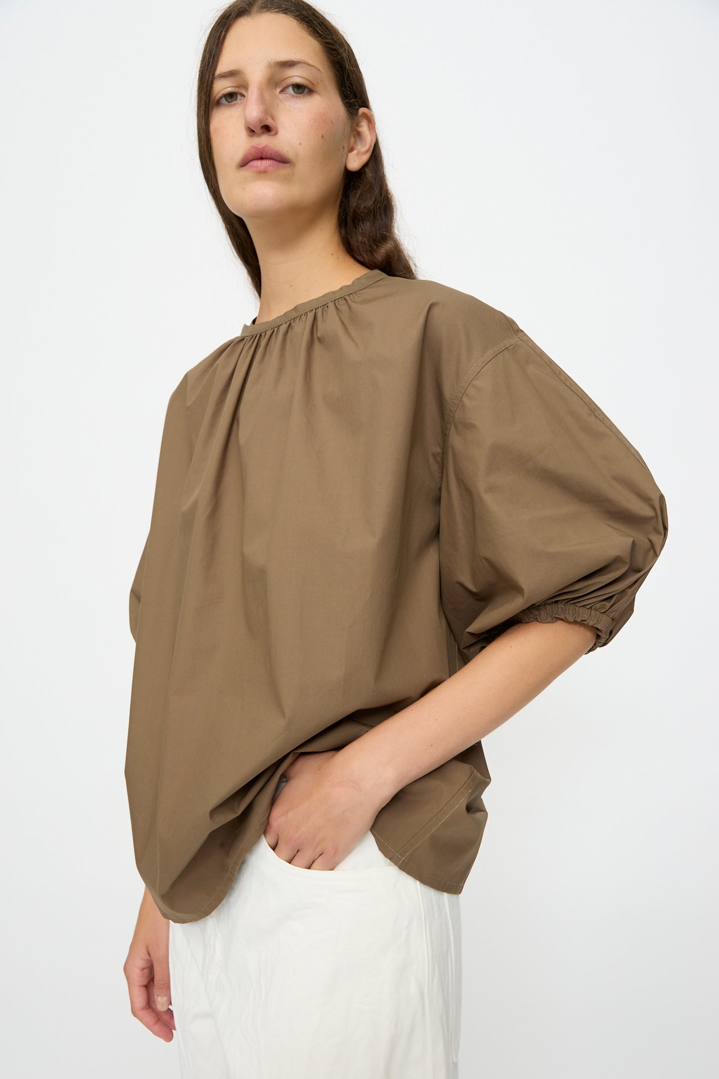 Someone wears the Studio Nicholson Cove Gathered Top in Boulder, a relaxed-fit brown top with bishop sleeves, paired with white pants against a plain light background.