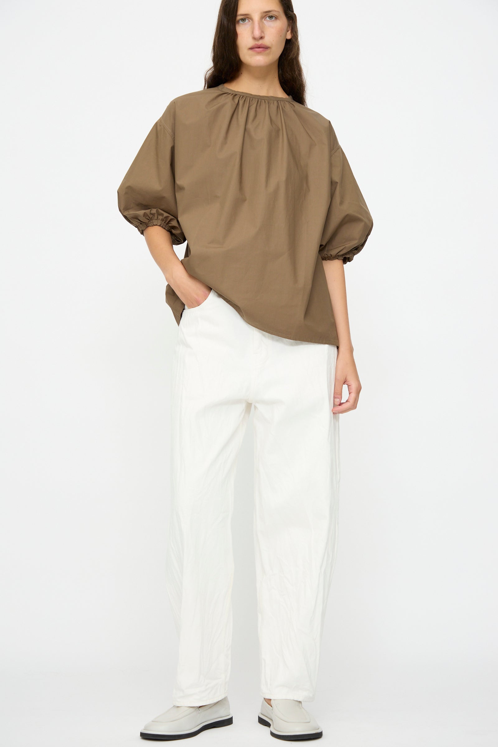 Person standing against a white background, wearing a loose brown top with gathered neckline, Studio Nicholson Denim Cahun Skater Pant in White, and light-colored shoes.