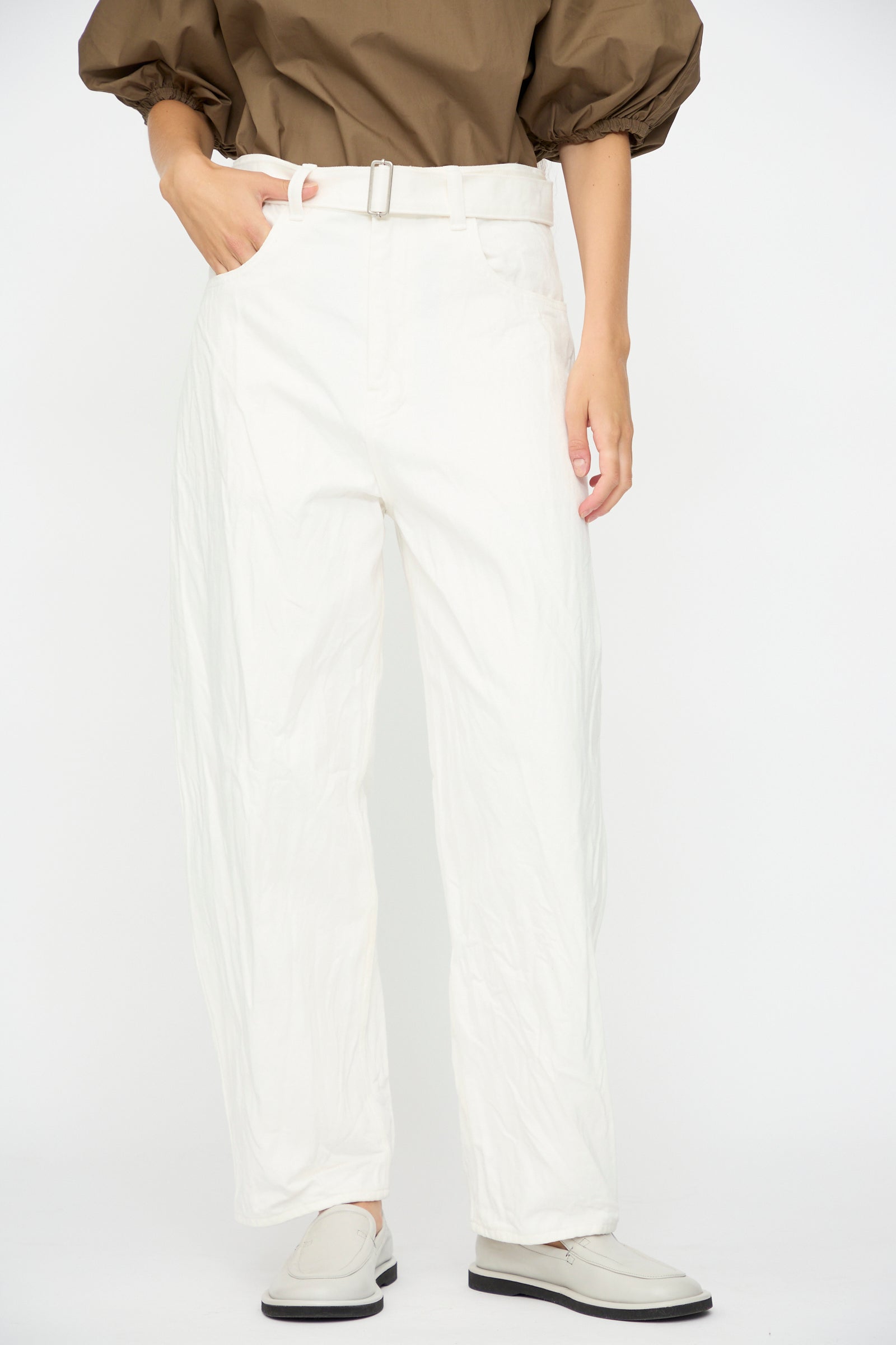 Person wearing a brown long-sleeve shirt and Studio Nicholson's Denim Cahun Skater Pant in White with a wide leg silhouette, paired with white loafers, standing against a white background.