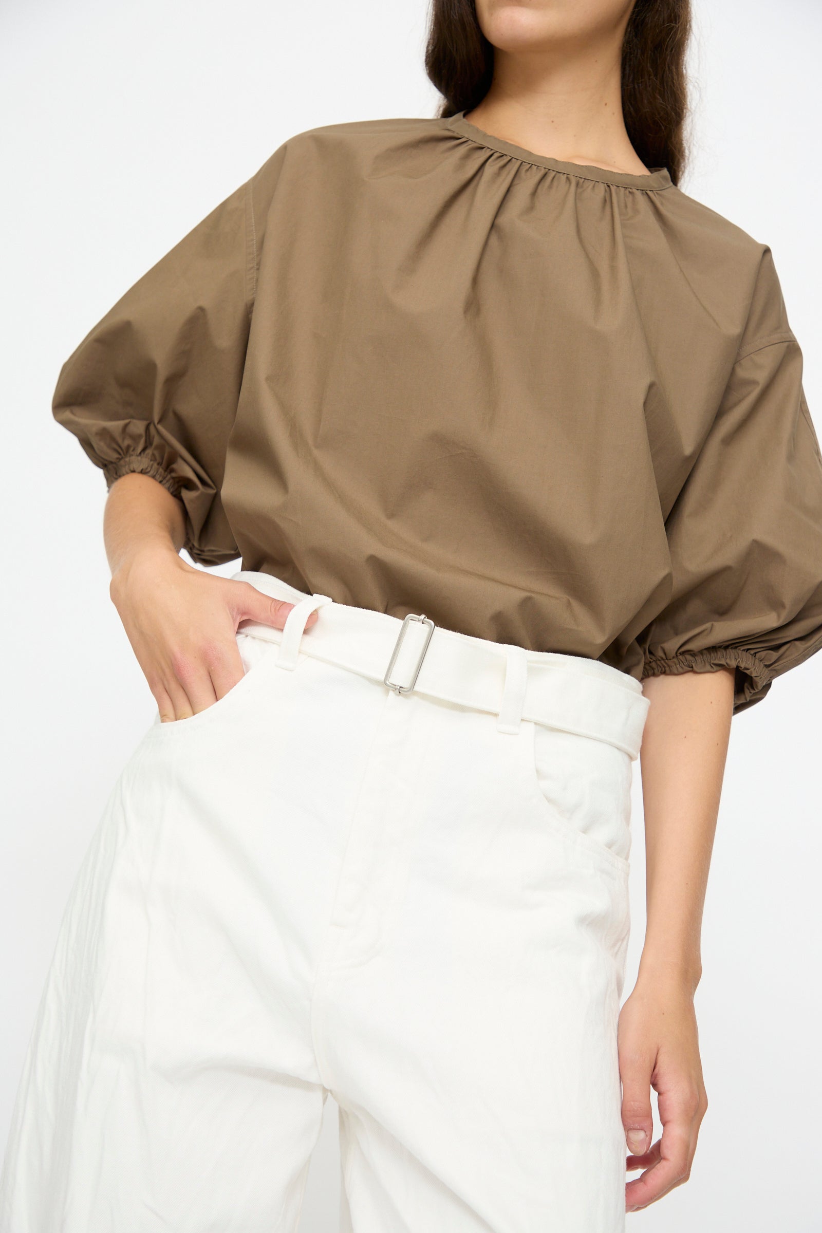 A person wears Studio Nicholson’s Denim Cahun Skater Pant in White—high waisted with a wide leg silhouette—paired with a loose brown blouse and white belt, standing against a plain background for a chic, contemporary look.