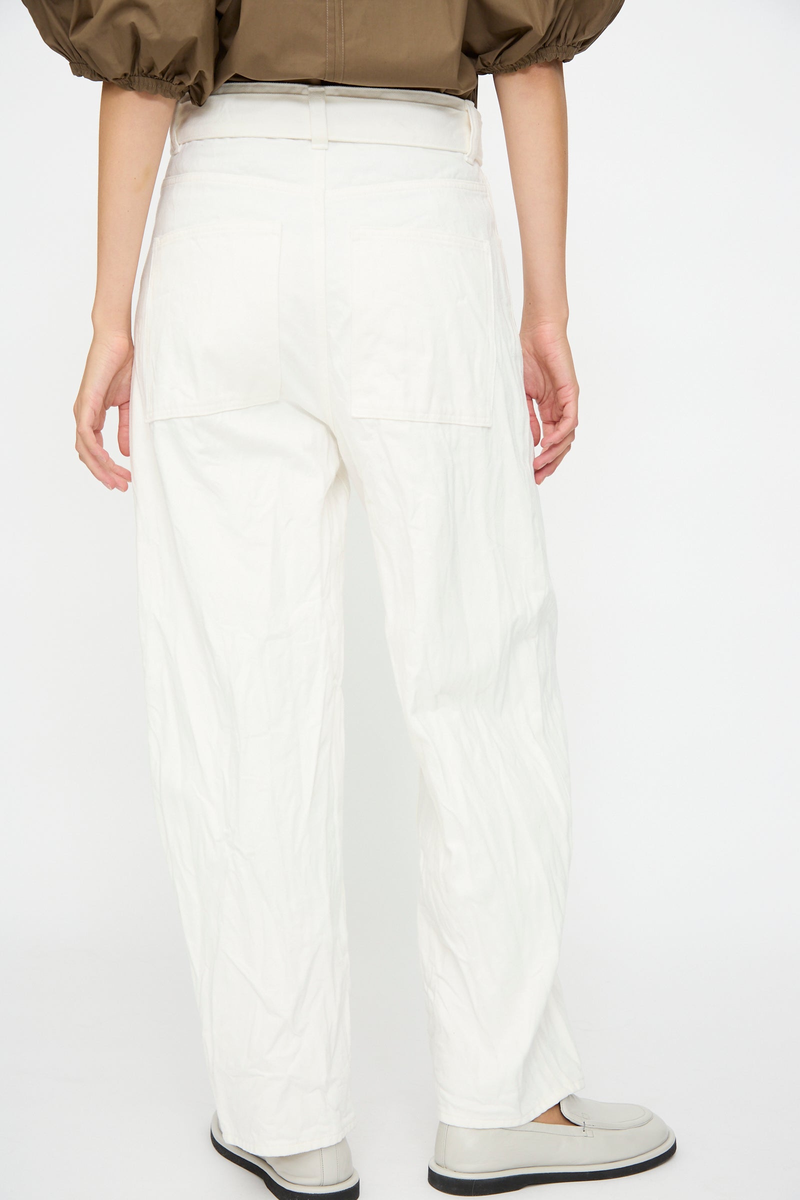 Person standing with their back to the camera, wearing Studio Nicholson's Denim Cahun Skater Pant in White, white shoes, and a brown puff sleeve top.