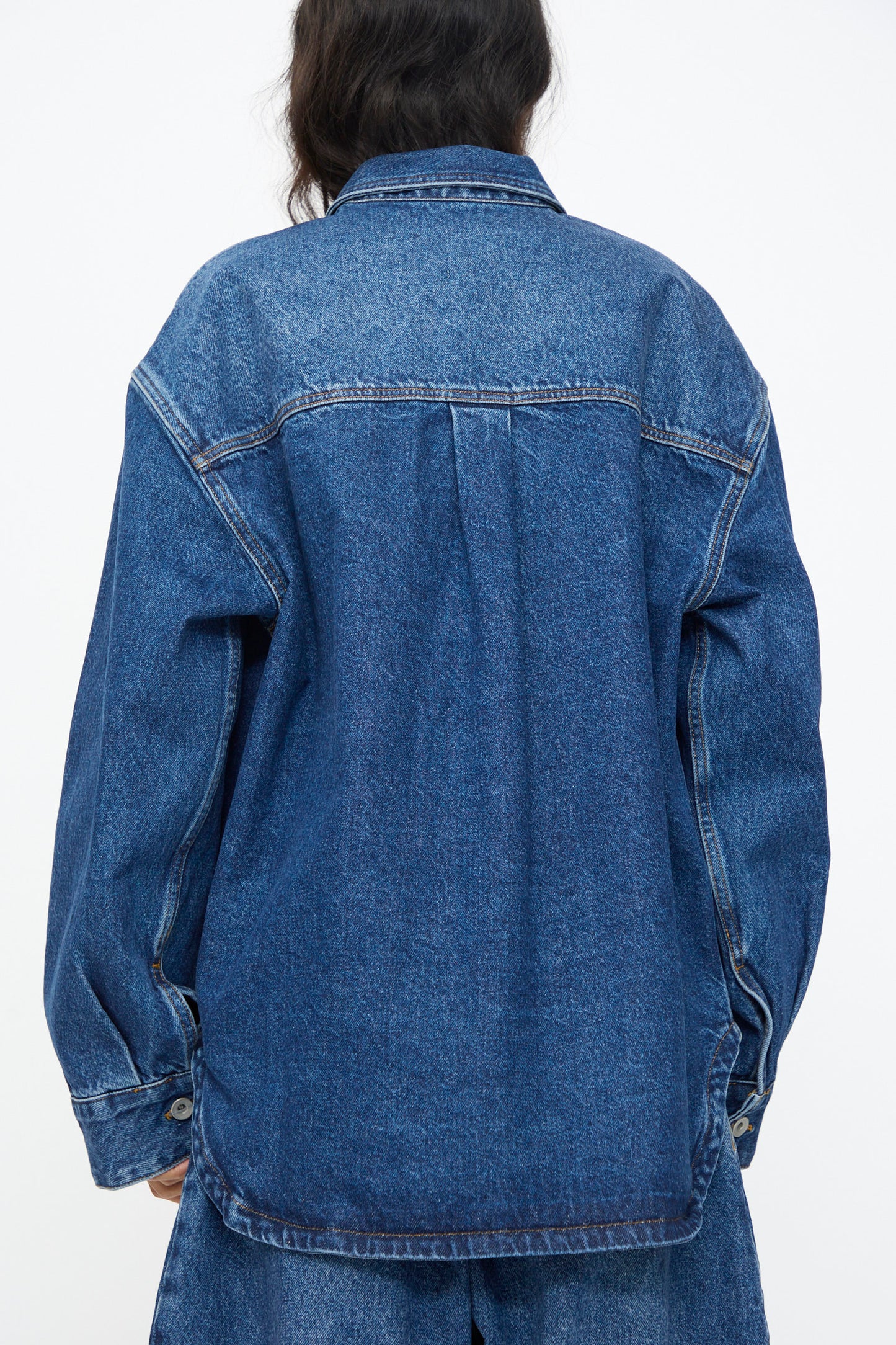 Model wears the Studio Nicholson Denim Paso Shirt in Worn Wash Indigo, a loose-fit style with oversized patch pockets, paired with matching denim bottoms. Shown from the back against a plain white background.