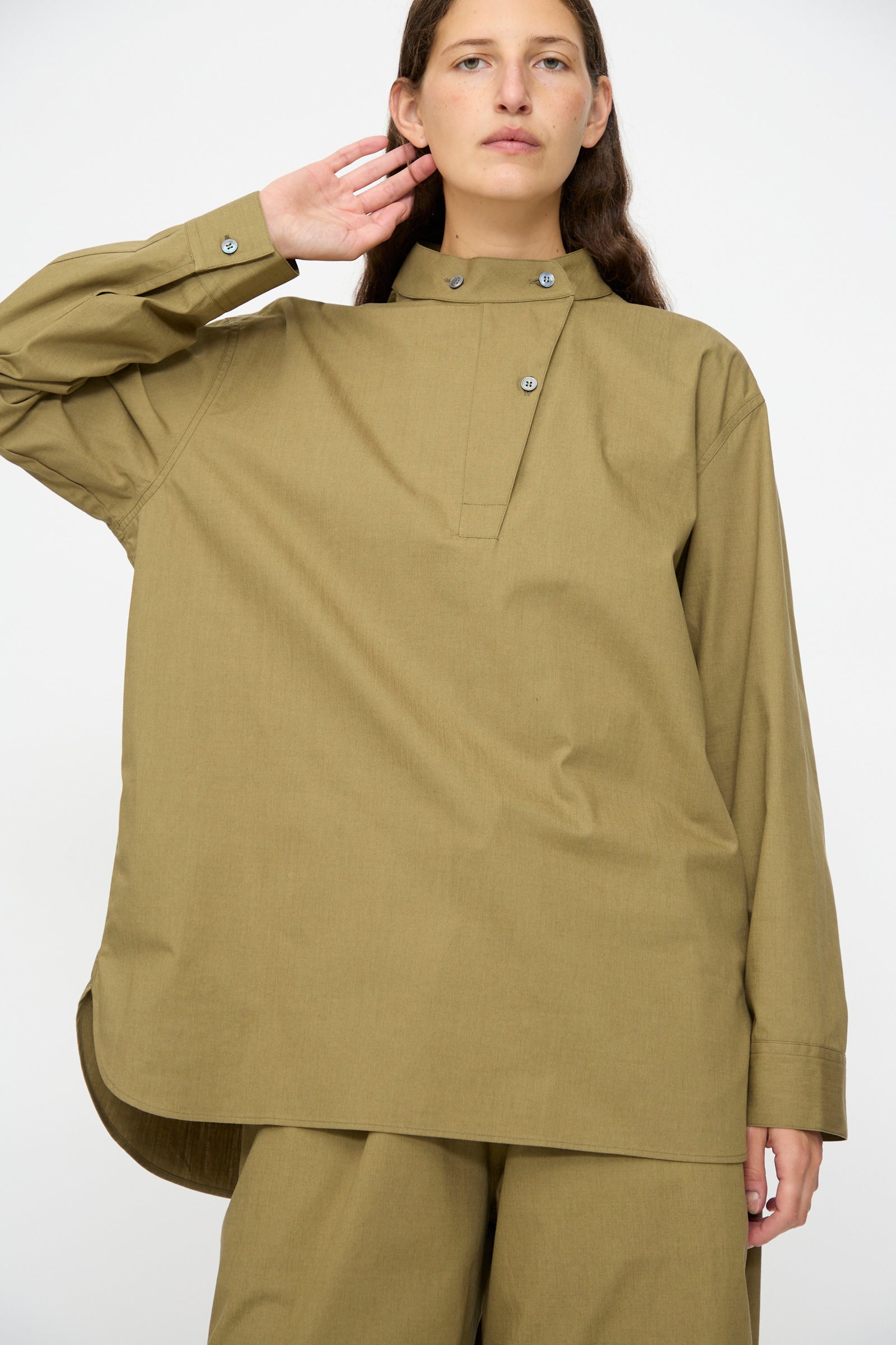 A person wears the Studio Nicholson Dimas Grandad Collar Shirt in Militaire with matching pants, standing against a plain white background, one hand raised near their face.