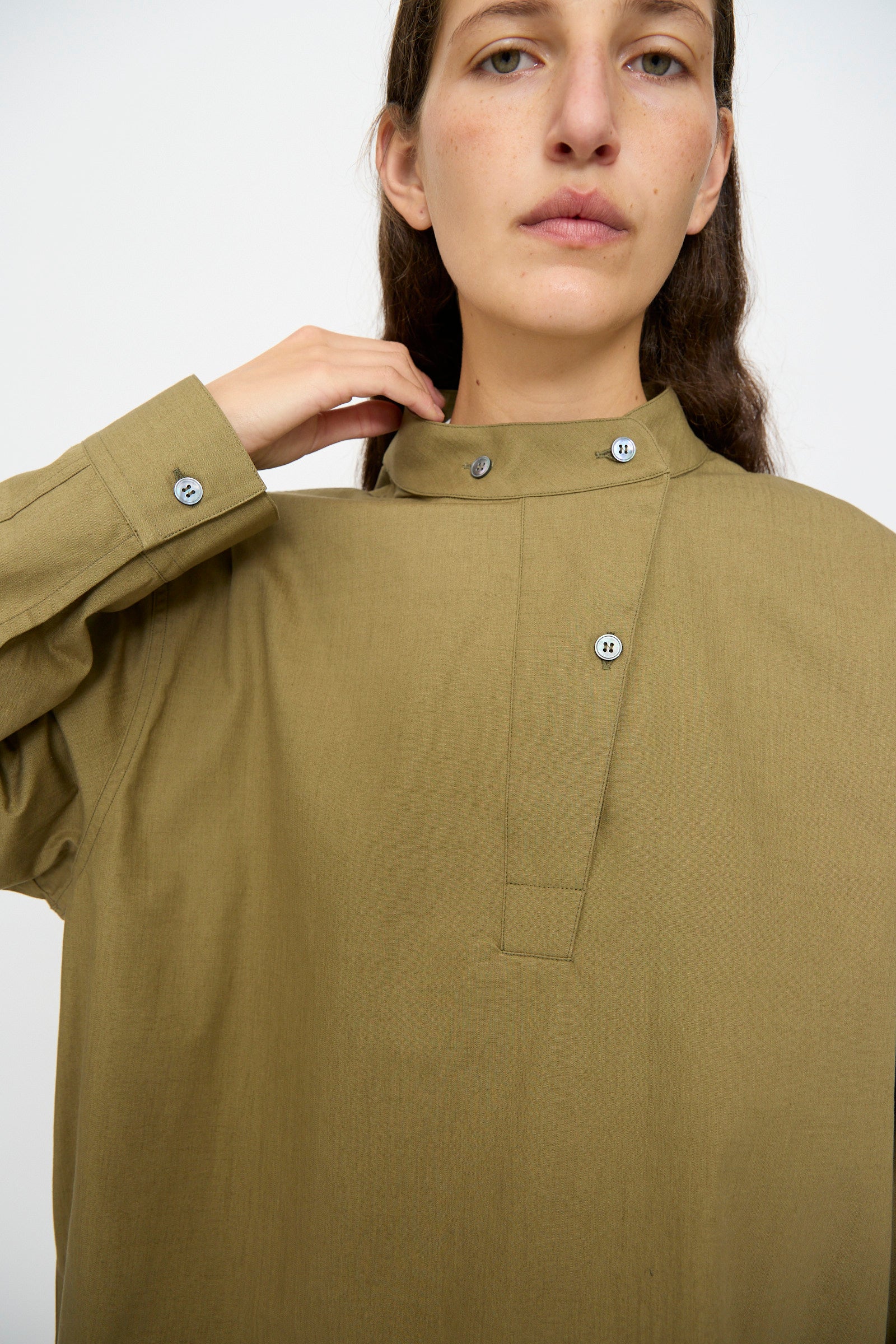 A person wearing the Studio Nicholson Dimas Grandad Collar Shirt in Militaire, an olive green vintage-style cotton long-sleeve with a high band collar, looks at the camera while touching the collar.