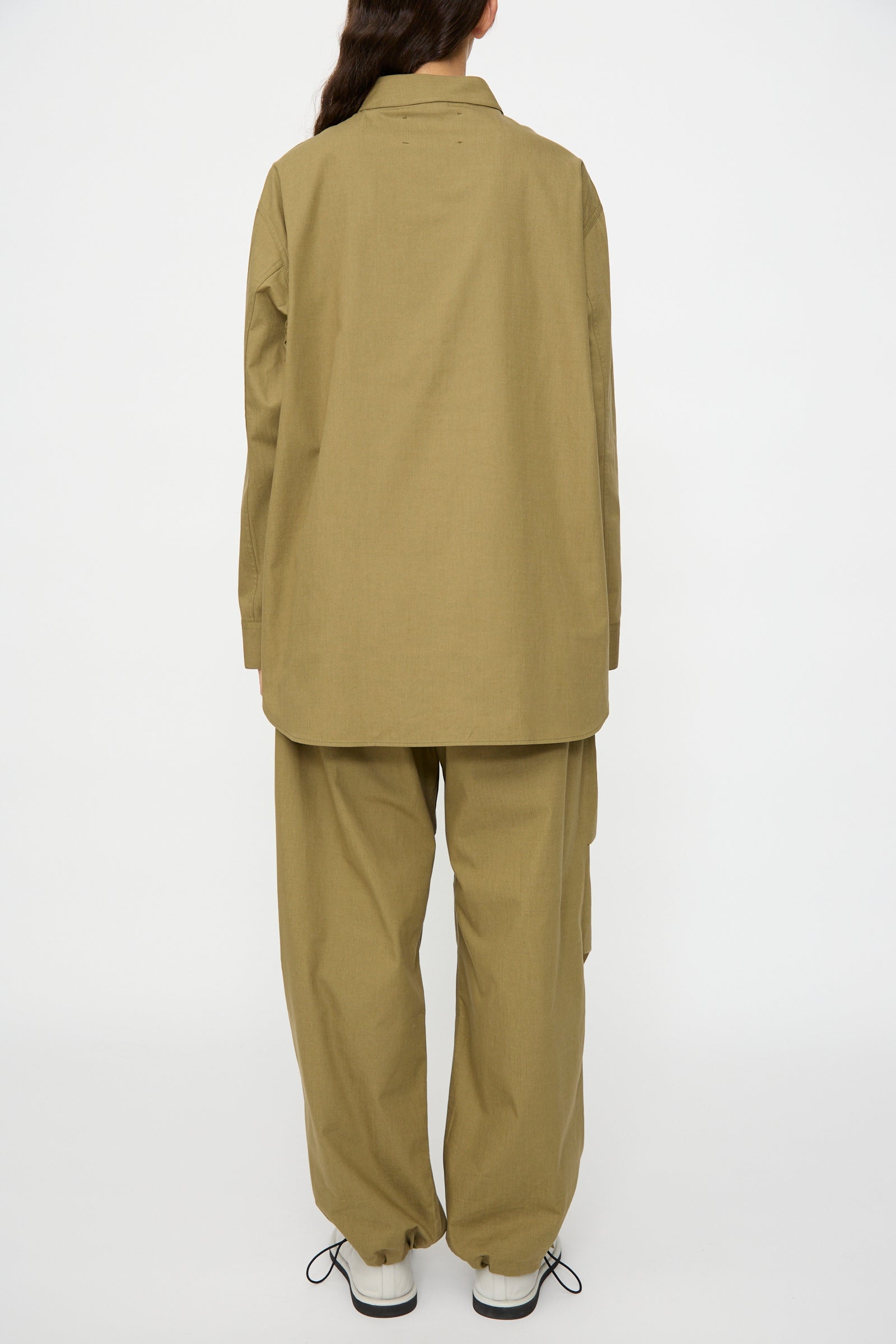 A person stands with their back to the camera, wearing Studio Nicholson’s olive Dimas Grandad Collar Shirt in Militaire and matching pants, paired with white shoes against a plain white background.