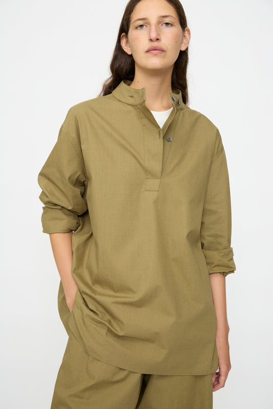 A person wears the Studio Nicholson Dimas Grandad Collar Shirt in Militaire with matching pants, hands in pockets, standing against a plain background.