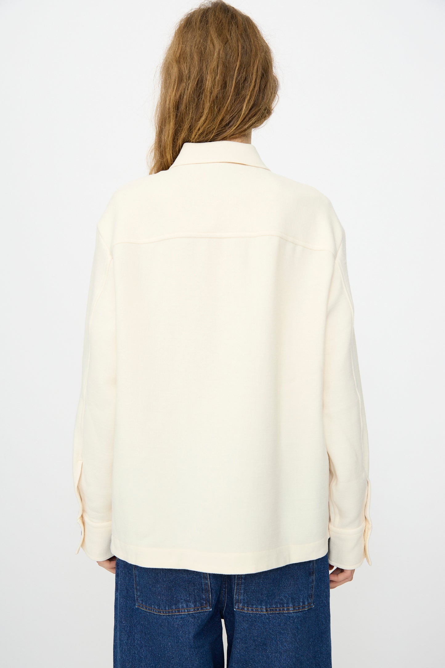 A person with long hair, seen from behind, wears the Studio Nicholson Double Wool Sablé Santini Overshirt in Chalk and blue jeans against a plain light background.