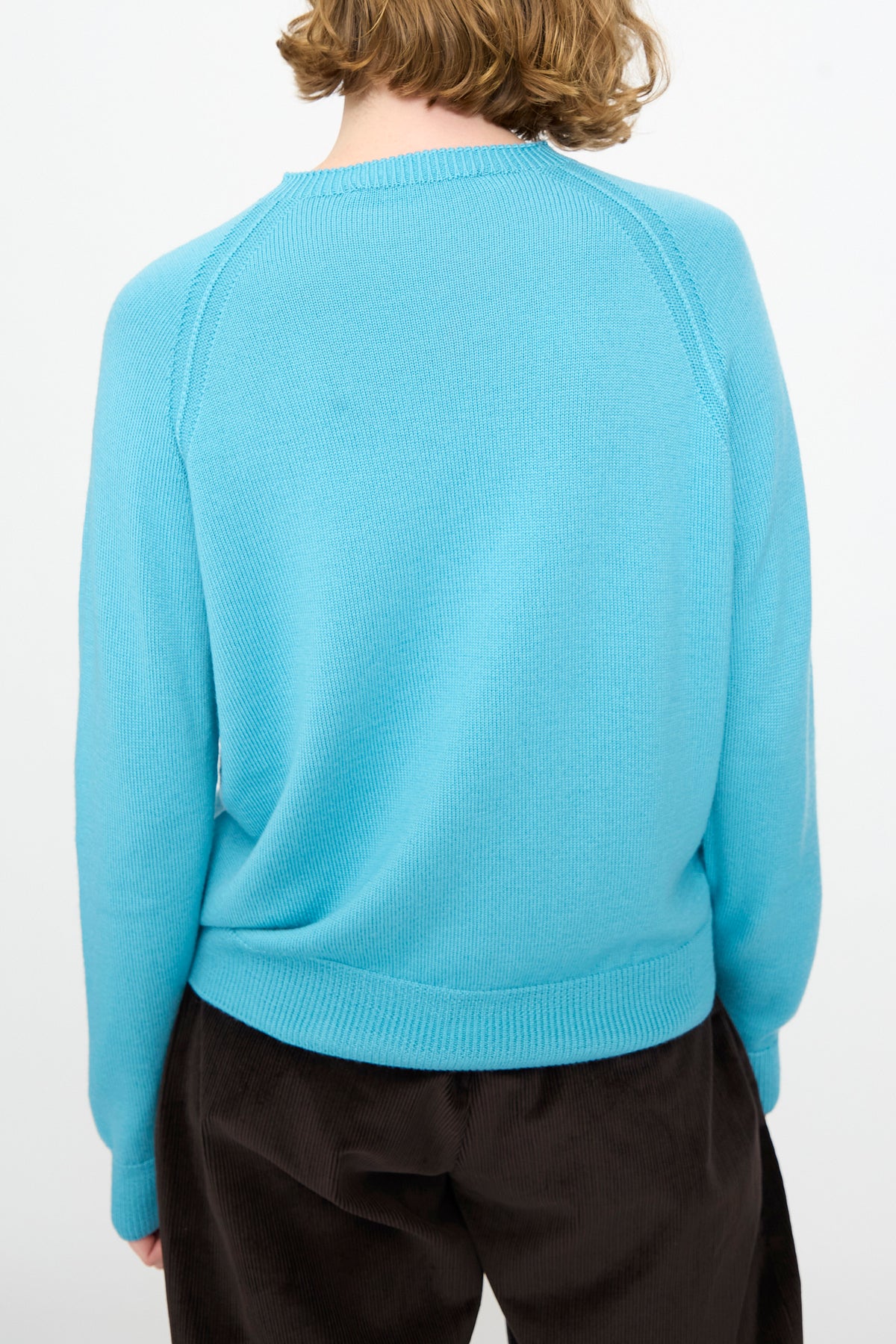 A person is shown from the back against a plain white background, wearing the Studio Nicholson Extrafine Merino Leonne Sweater in Sky and dark pants.