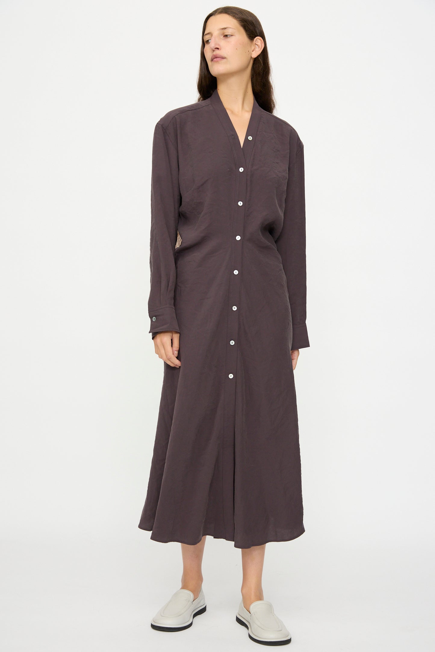 A person wears the Studio Nicholson Fiore Shirt Dress in Yam—an Italian-milled modal, long-sleeved button-down with white buttons—and light slip-on shoes, standing against a plain white background.