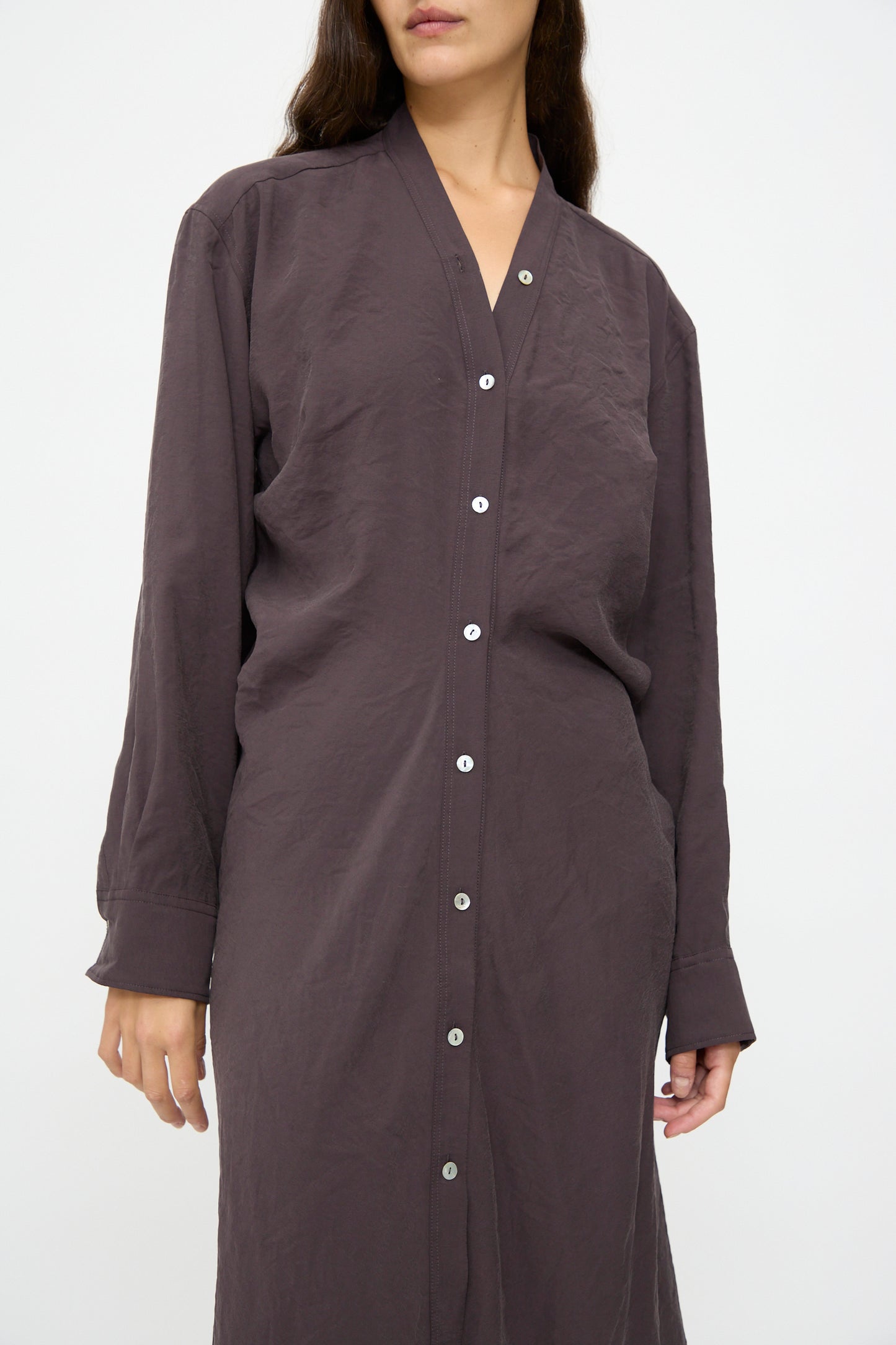 A woman stands against a plain white background wearing the Studio Nicholson Fiore Shirt Dress in Yam, a long-sleeve, button-down piece with a V-neck collar crafted from Italian-milled modal.