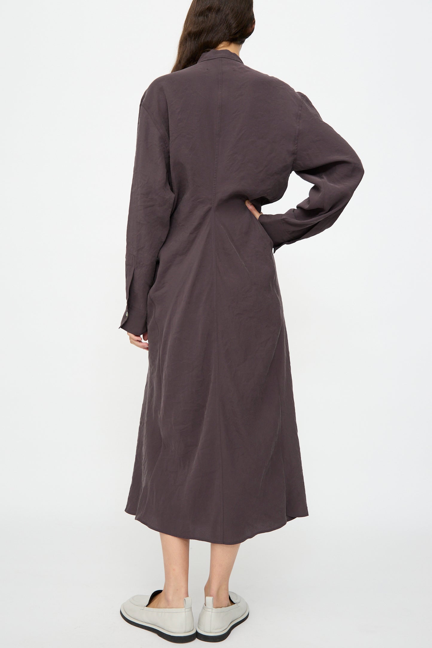 A person stands facing away against a white background, wearing the Studio Nicholson Fiore Shirt Dress in Yam with long sleeves and white slip-on shoes.