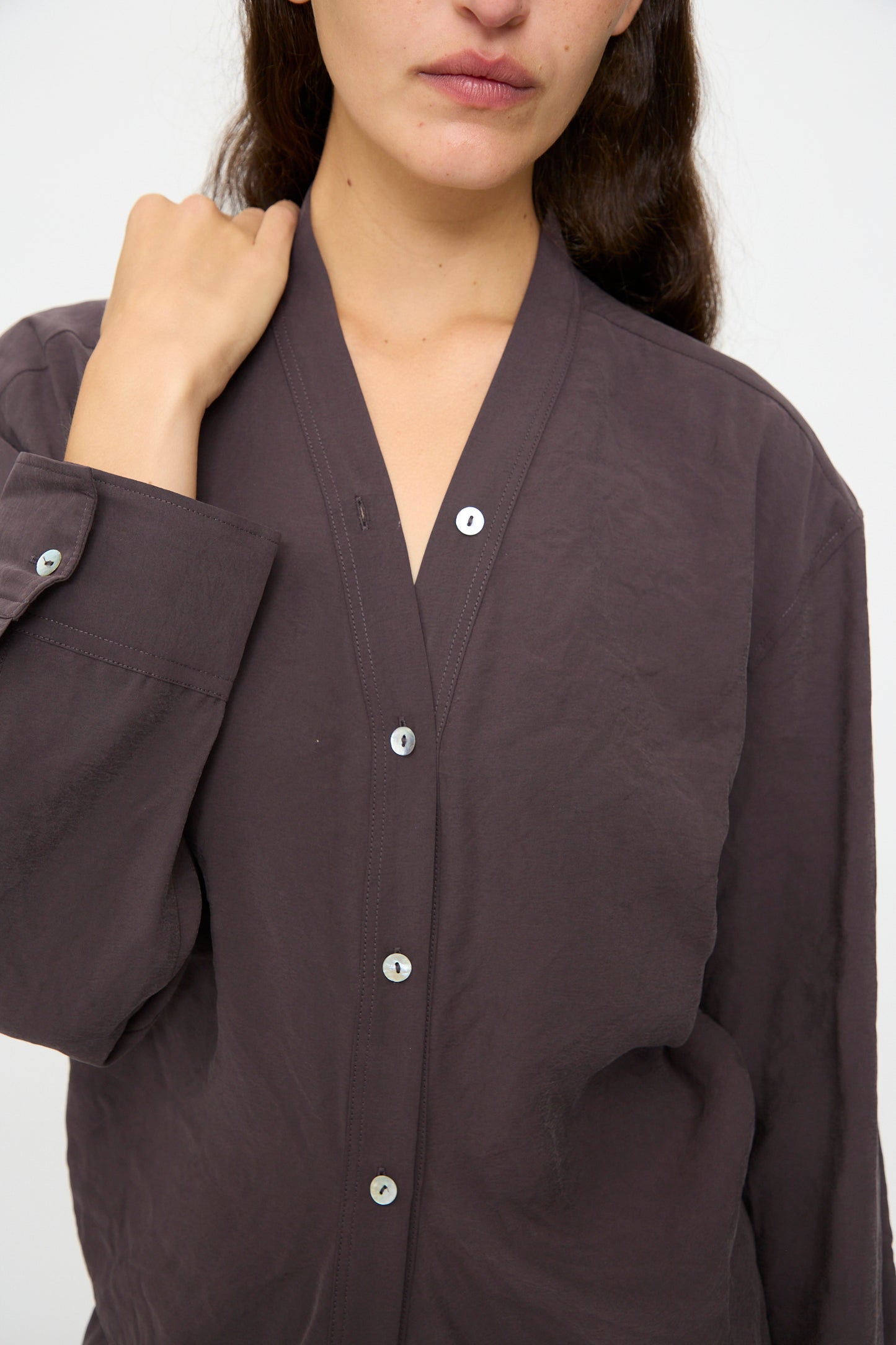 A person wearing the Studio Nicholson Fiore Shirt Dress in Yam, a loose long-sleeve button-up, stands against a plain background with one hand touching the collar.
