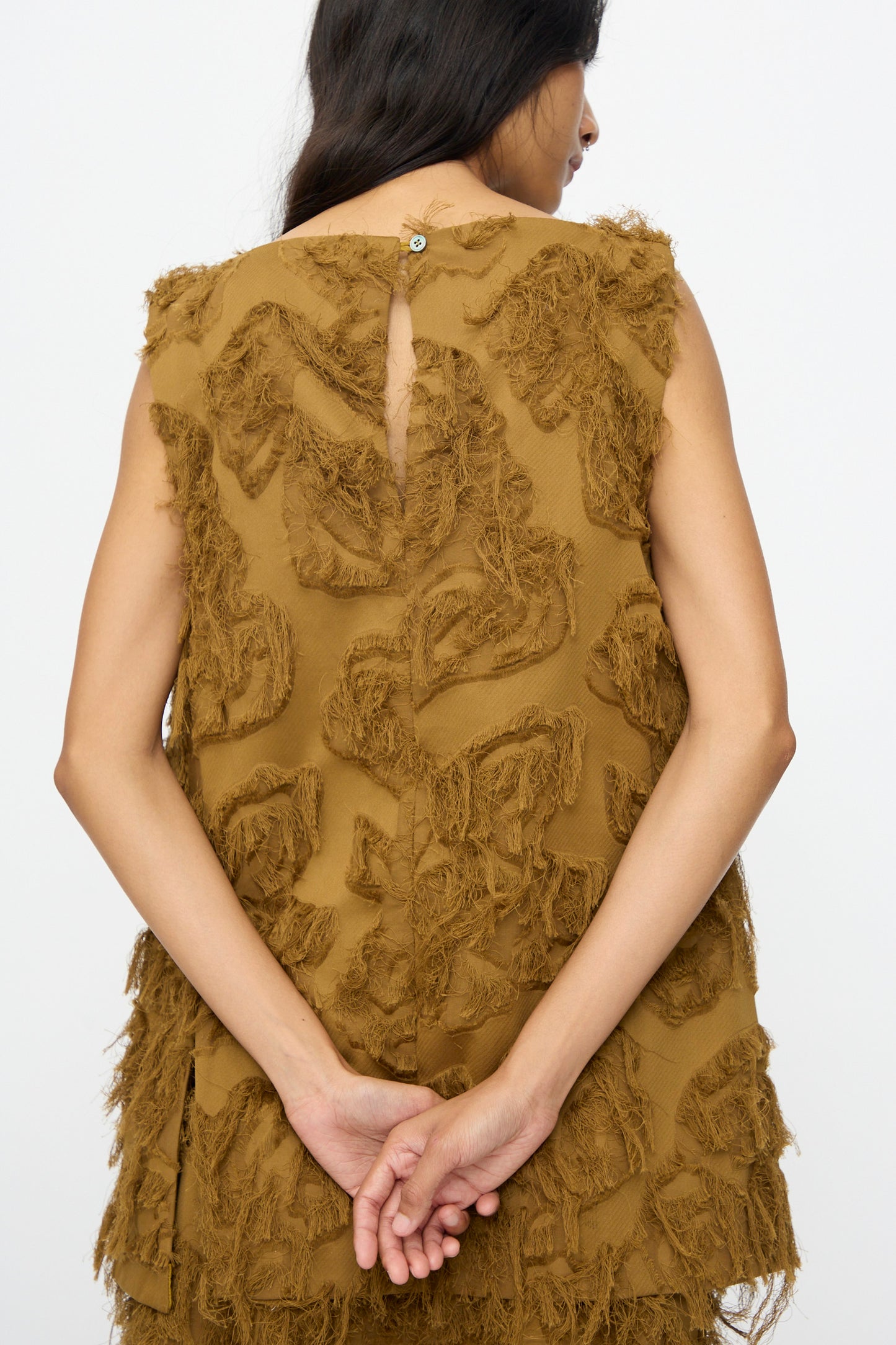 A person with long dark hair, wearing the Studio Nicholson Fringe Jacquard Mager Top in Mustard, stands with their back to the camera and hands clasped behind them.