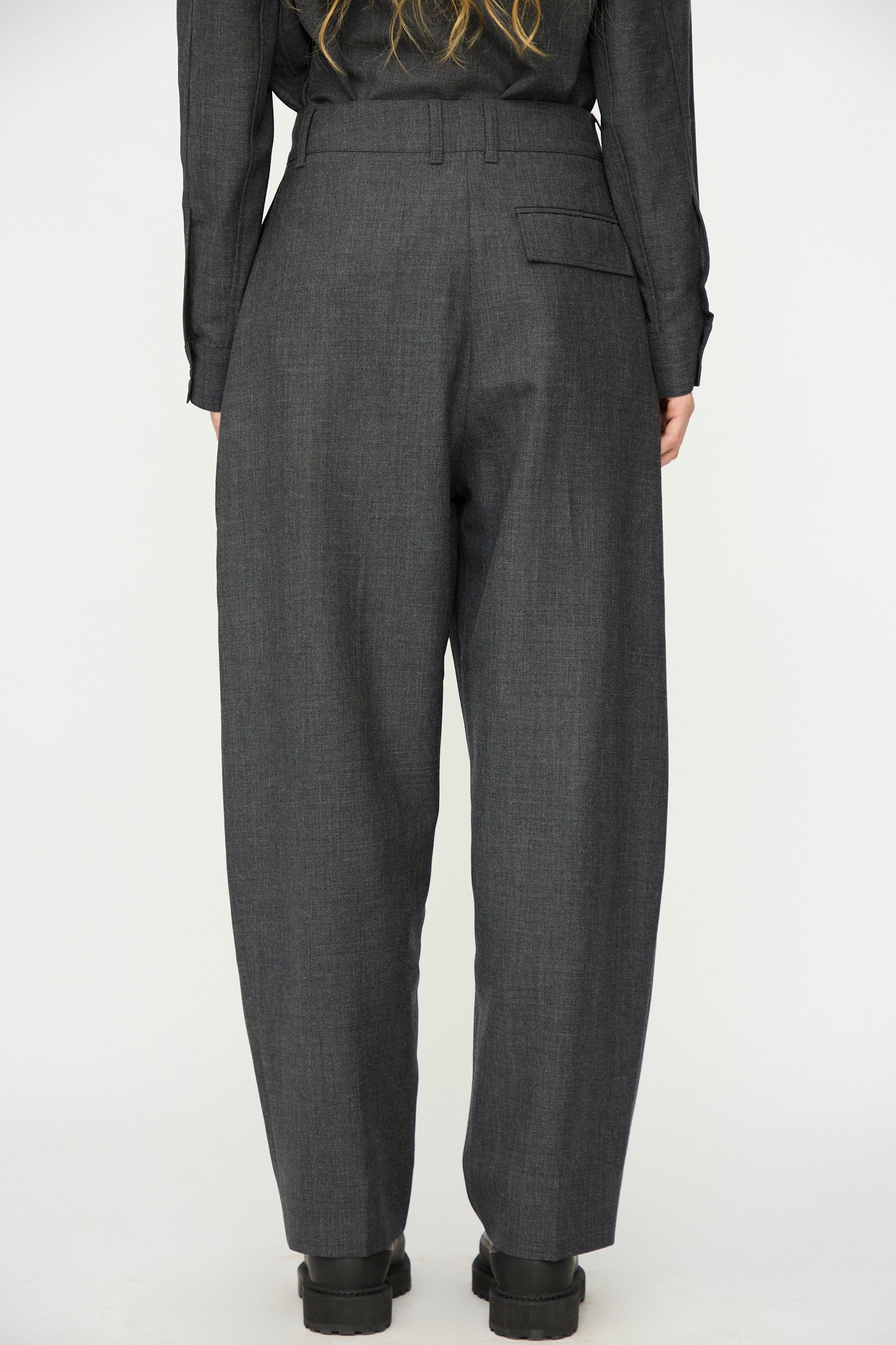 A person wearing Studio Nicholson's Heritage Wool Acuna Pant in Slate Melange and black shoes stands with their back to the camera against a plain background.