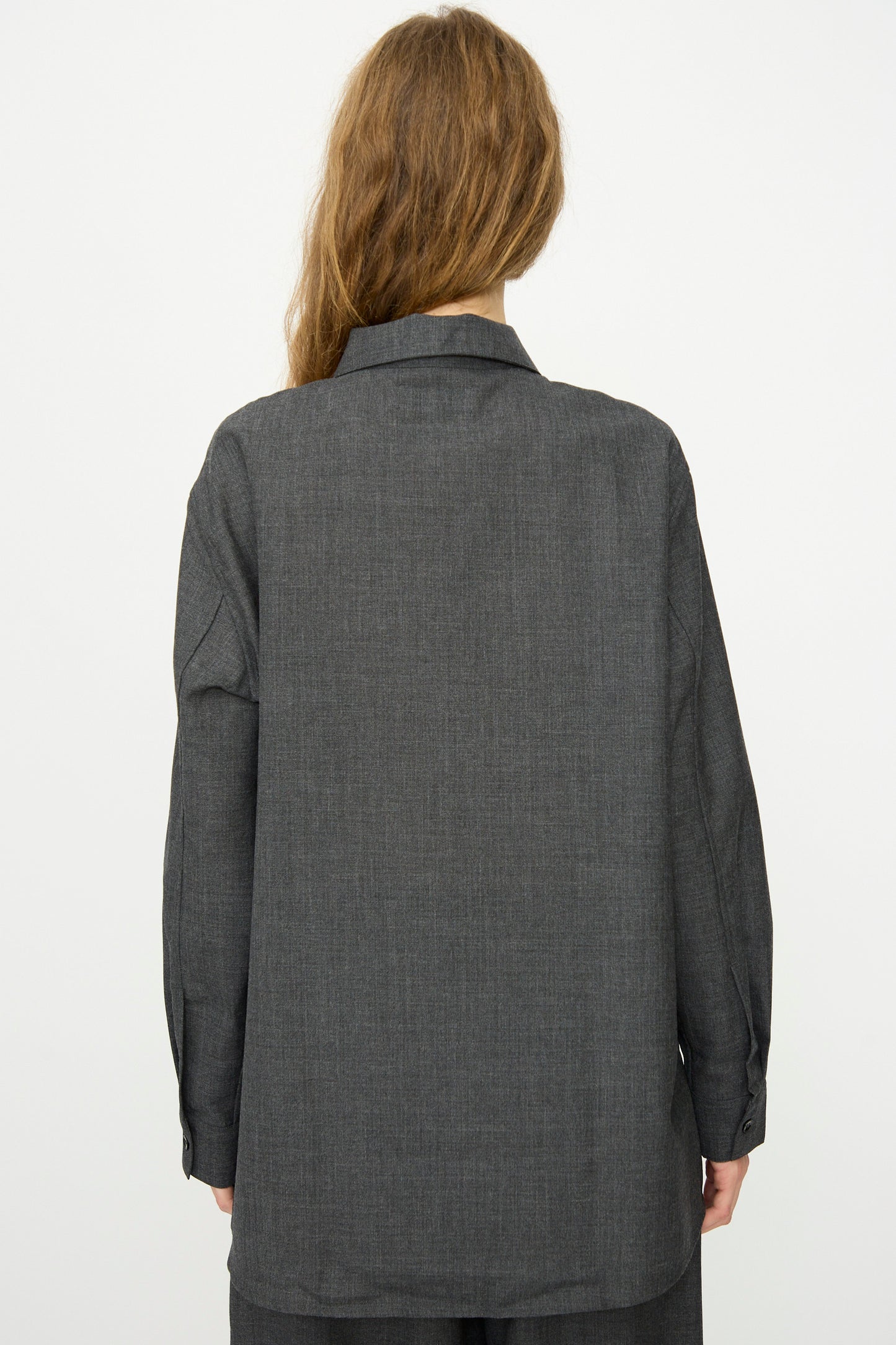 A person with long, light brown hair is seen from the back wearing Studio Nicholson's Heritage Wool Pinole Shirt in Slate Melange, a relaxed-fit dark gray long sleeve, against a plain light background.