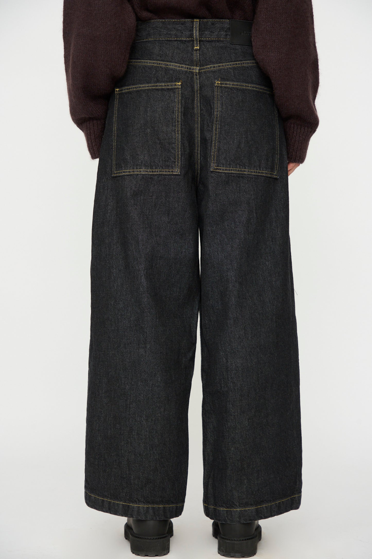 Model wears Studio Nicholson's Italian Denim Neive Pant in Black, featuring wide legs and large back pockets, paired with a dark sweater. Shown from the back against a plain light background.