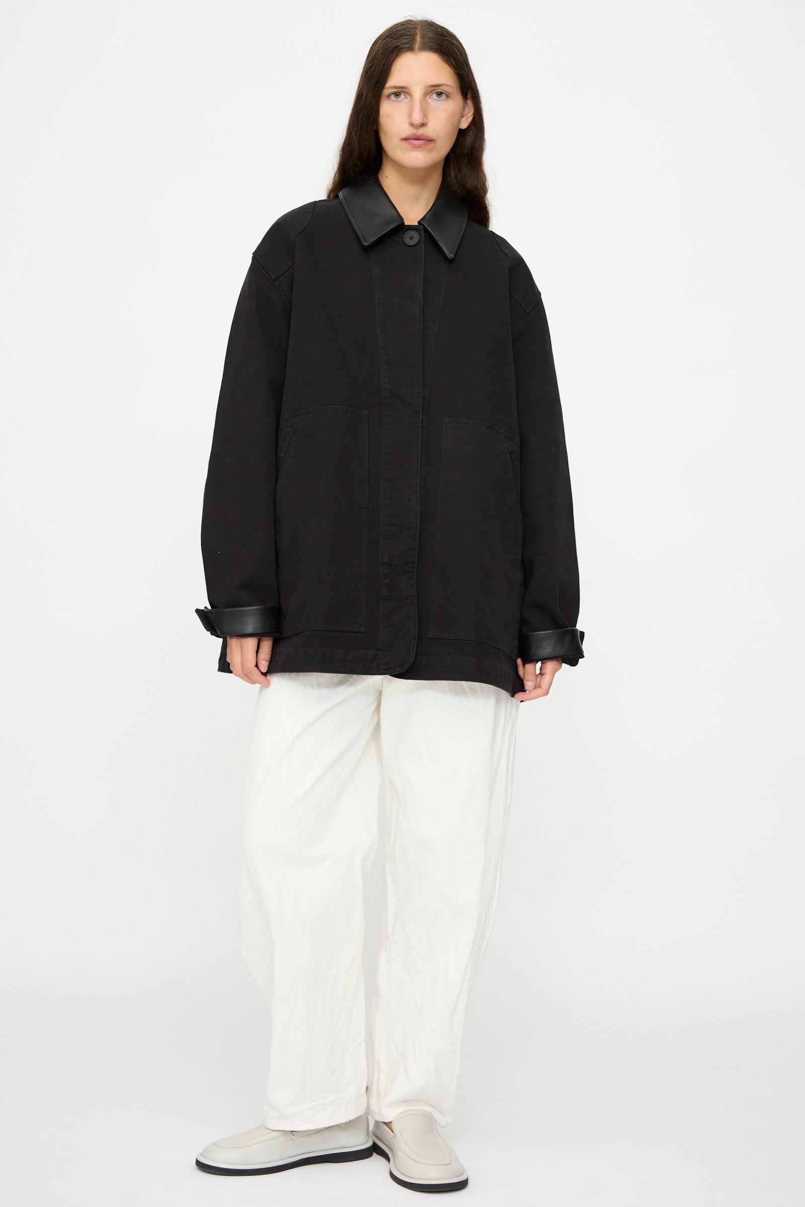 A person stands against a plain background wearing the Studio Nicholson Leader Chore Jacket in Darkest Navy, styled with white wide-leg pants and white slip-on shoes.