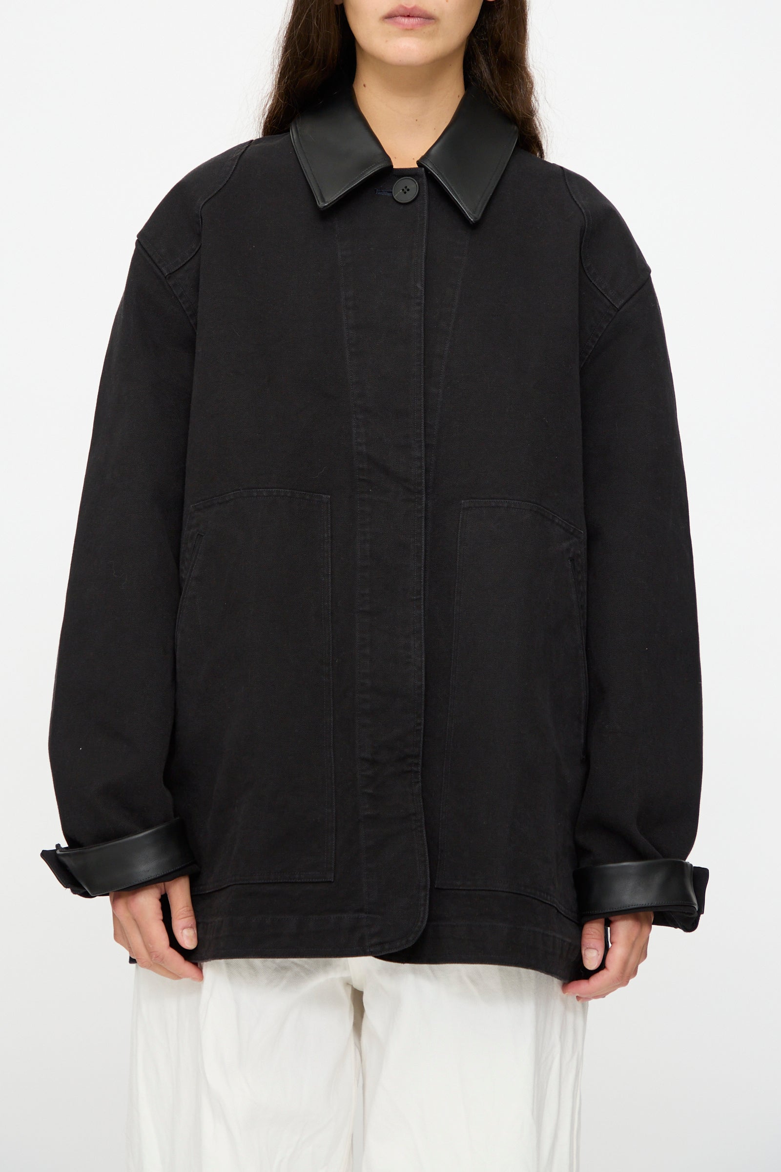 A person wears the Studio Nicholson Leader Chore Jacket in Darkest Navy—relaxed fit, heavyweight canvas with a leather collar and cuffs—paired with loose white pants, standing against a plain white background.