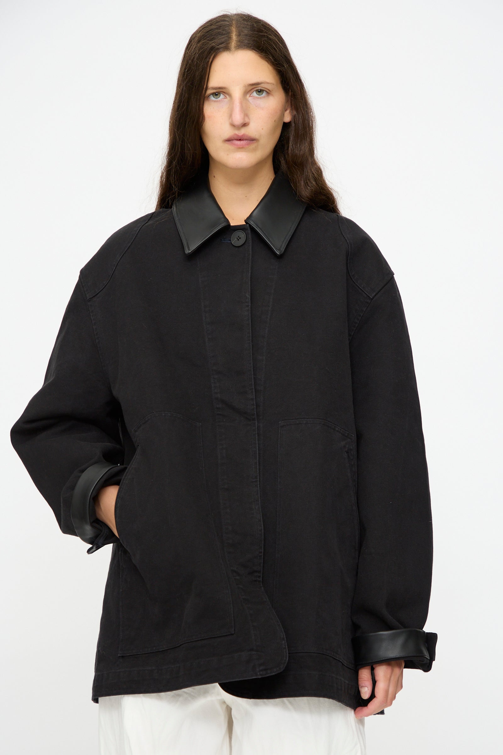 A person with long brown hair wears Studio Nicholson's Leader Chore Jacket in Darkest Navy, featuring a relaxed, oversized fit with a leather collar and cuffs, styled over white pants against a plain white background.