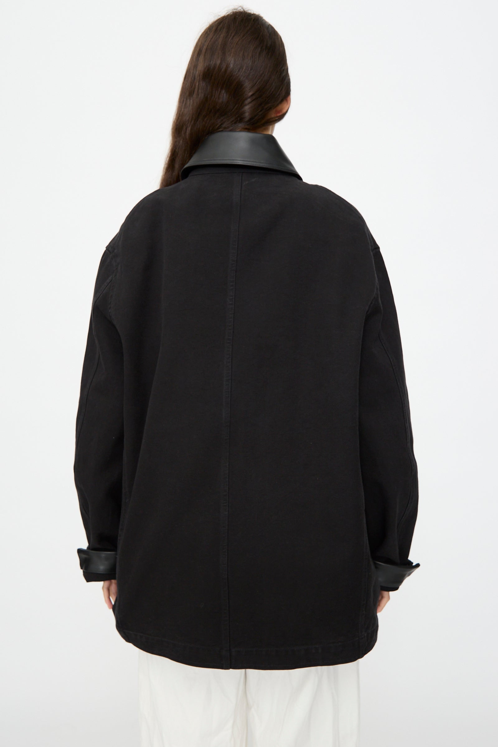 Person with long brown hair shown from the back, wearing Studio Nicholson's Leader Chore Jacket in Darkest Navy—an oversized, relaxed fit with a wide collar—paired with white pants against a plain background.