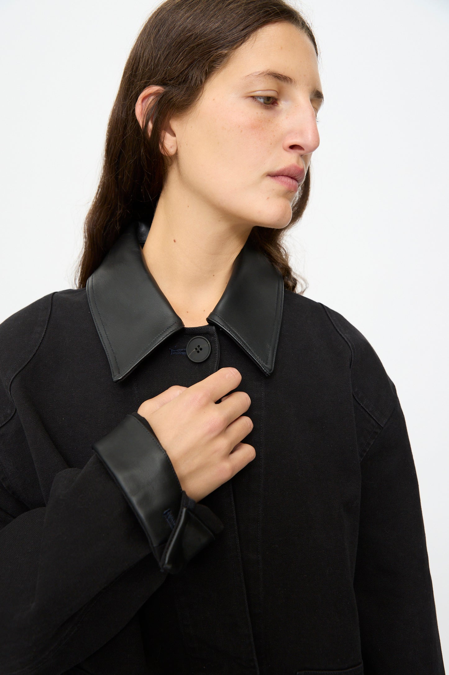 A person with long brown hair wears the Studio Nicholson Leader Chore Jacket in Darkest Navy, a relaxed fit coat with a wide leather collar and cuffs, gazing to the side against a plain background.