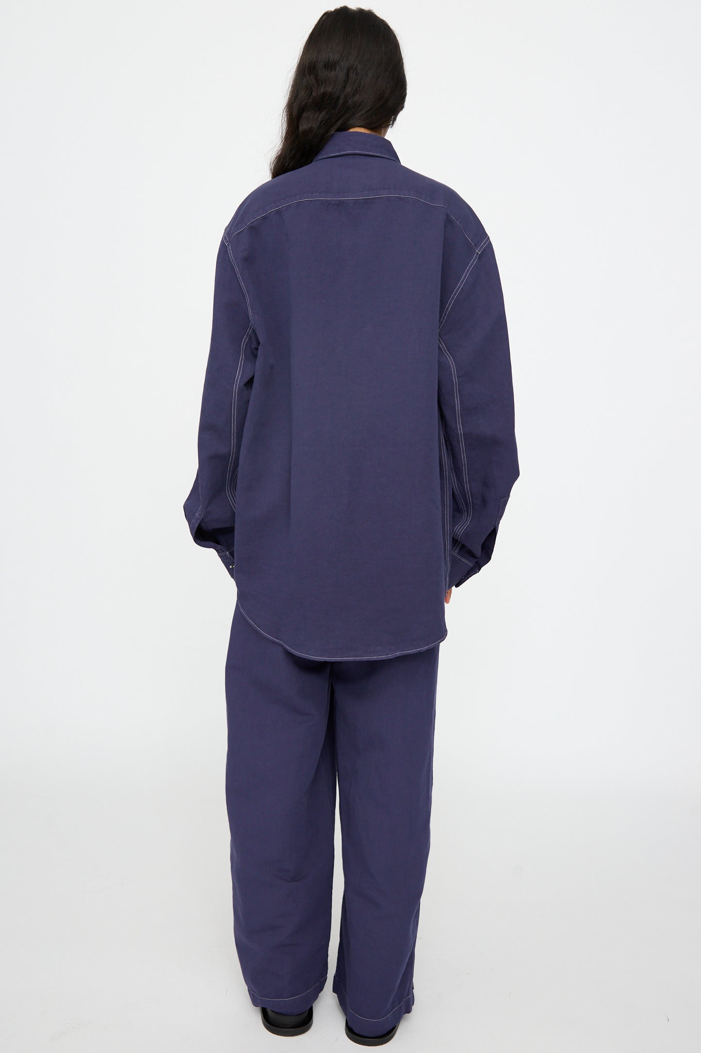 A person stands with their back to the camera, wearing the Studio Nicholson Louro Utility Pocket Shirt in Indigo and matching pants against a plain white background.