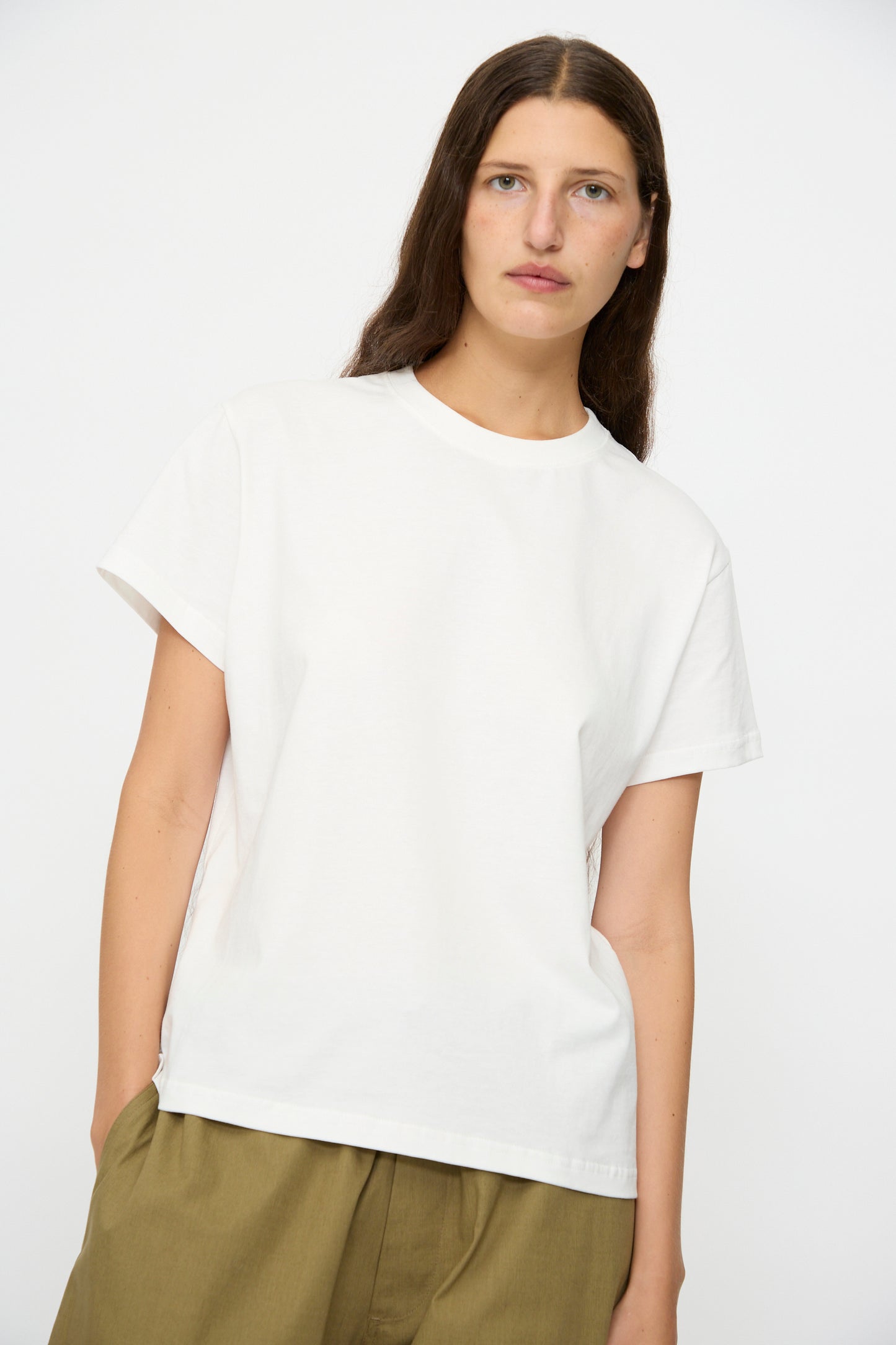 A person with long brown hair wears the Studio Nicholson Marine Perfect Tee in Off White and olive green pants, standing against a plain white background.