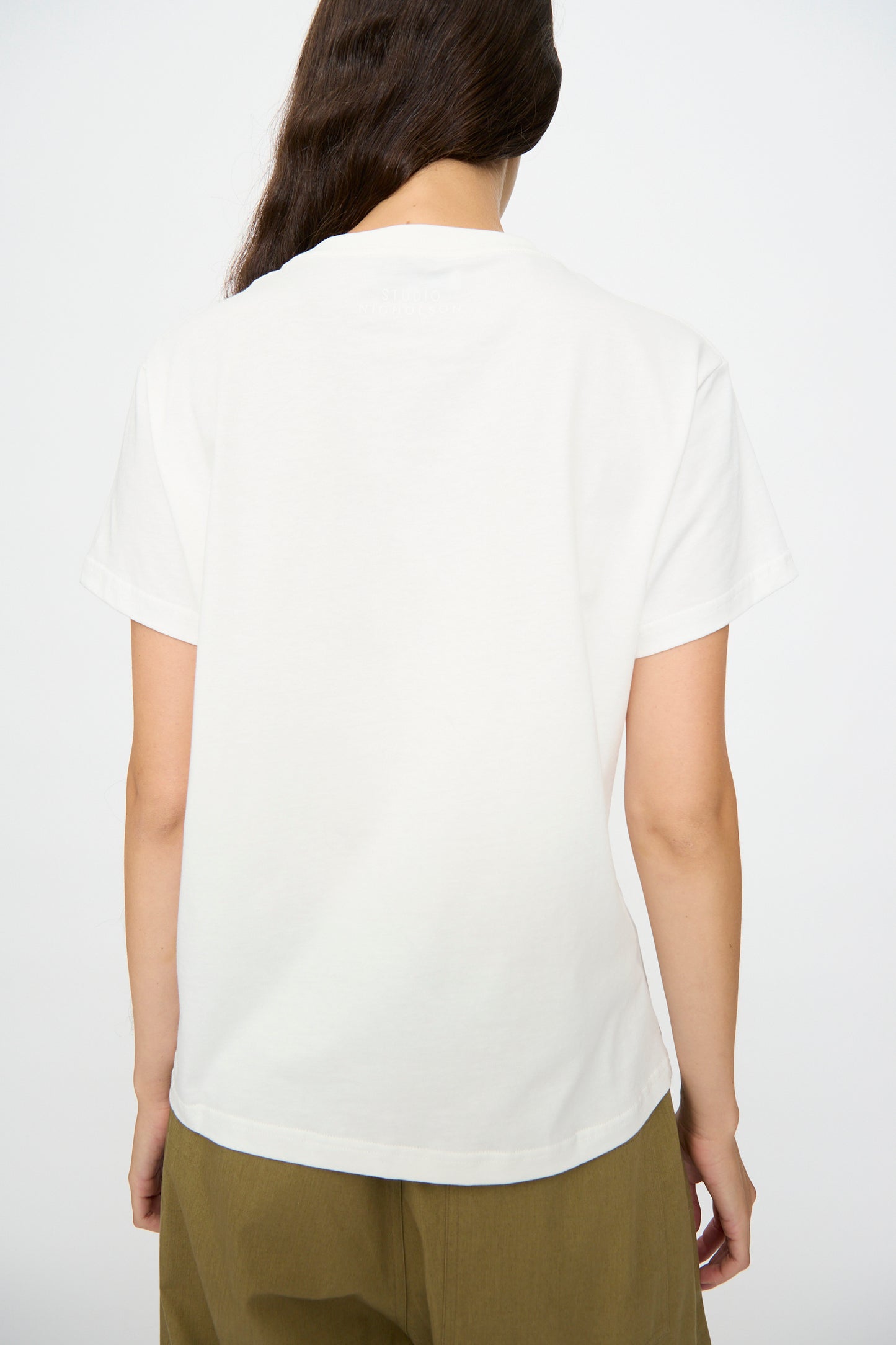 A person with long brown hair is shown from the back, wearing Studio Nicholson’s Marine Perfect Tee in Off White and olive green pants, set against a plain background.